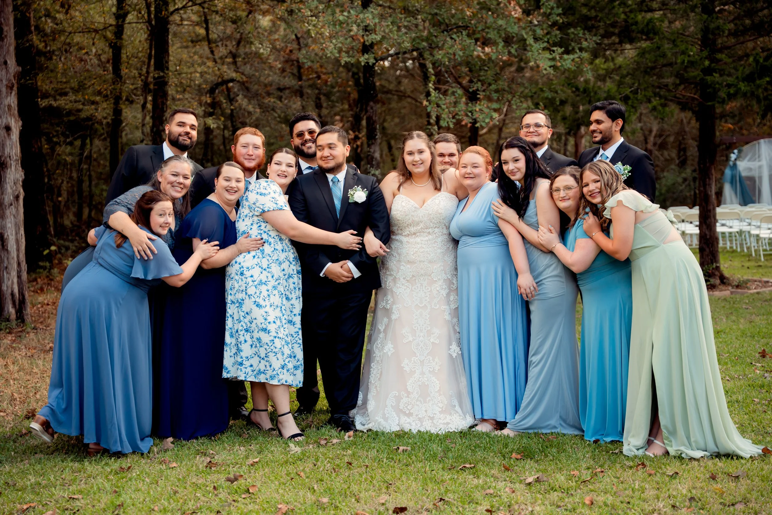 A group of people at a wedding outdoors in a wooded area. The bride and groom are in the center, the bride in a white lace wedding dress and the groom in a black suit. Family and friends surround them, many wearing blue or pastel-colored dresses and 