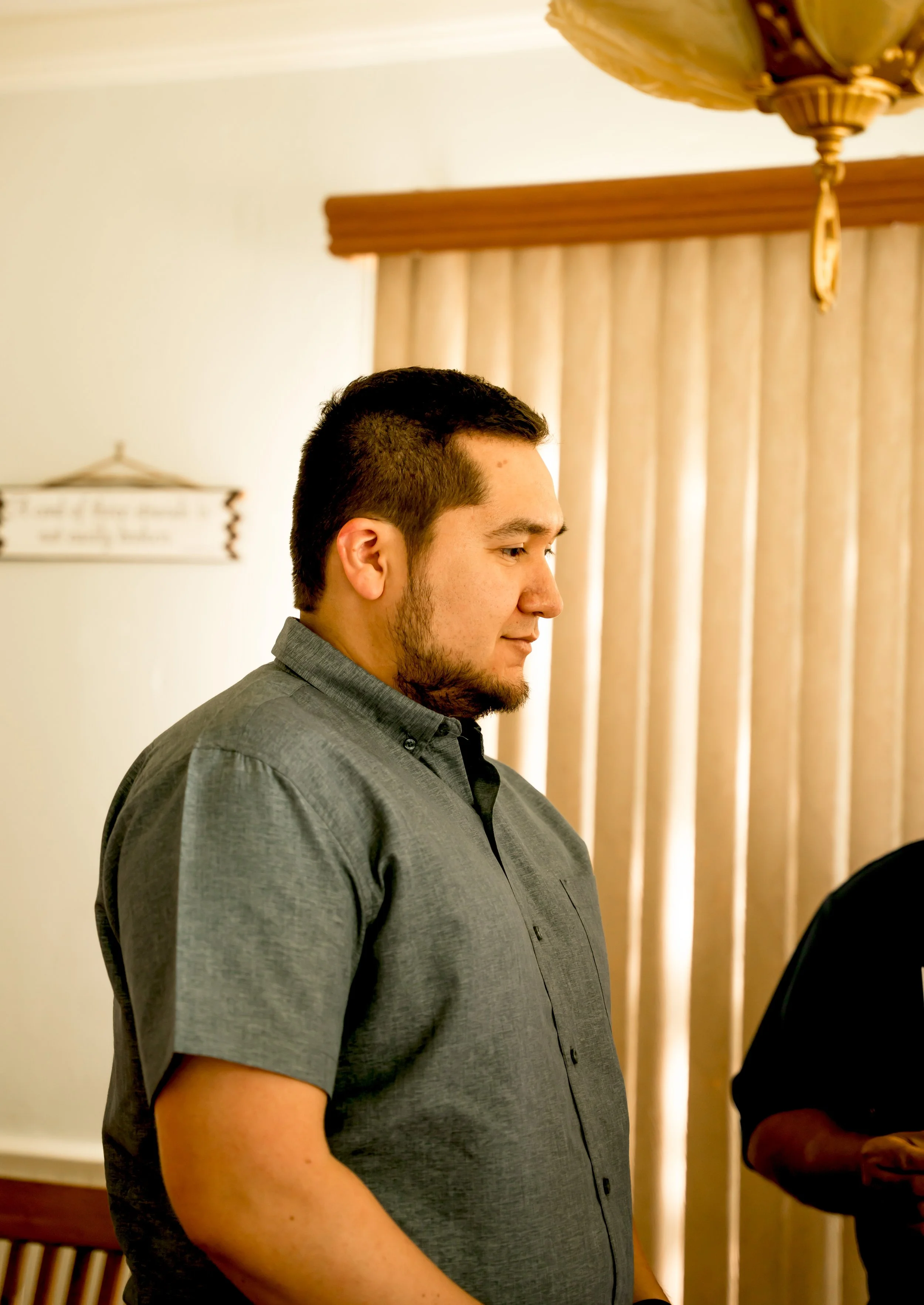 Profile of a young man with short dark hair, wearing a gray button-up shirt, standing indoors with beige curtains in the background.