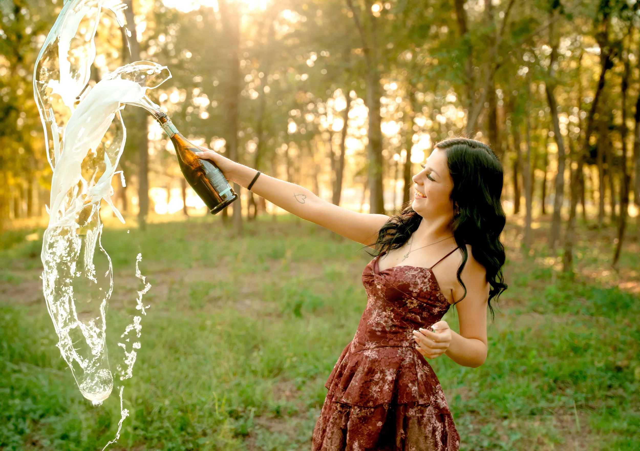 A woman outdoors in a wooded area, smiling, holding a bottle and pouring beer into a glass, with beer spilling out.