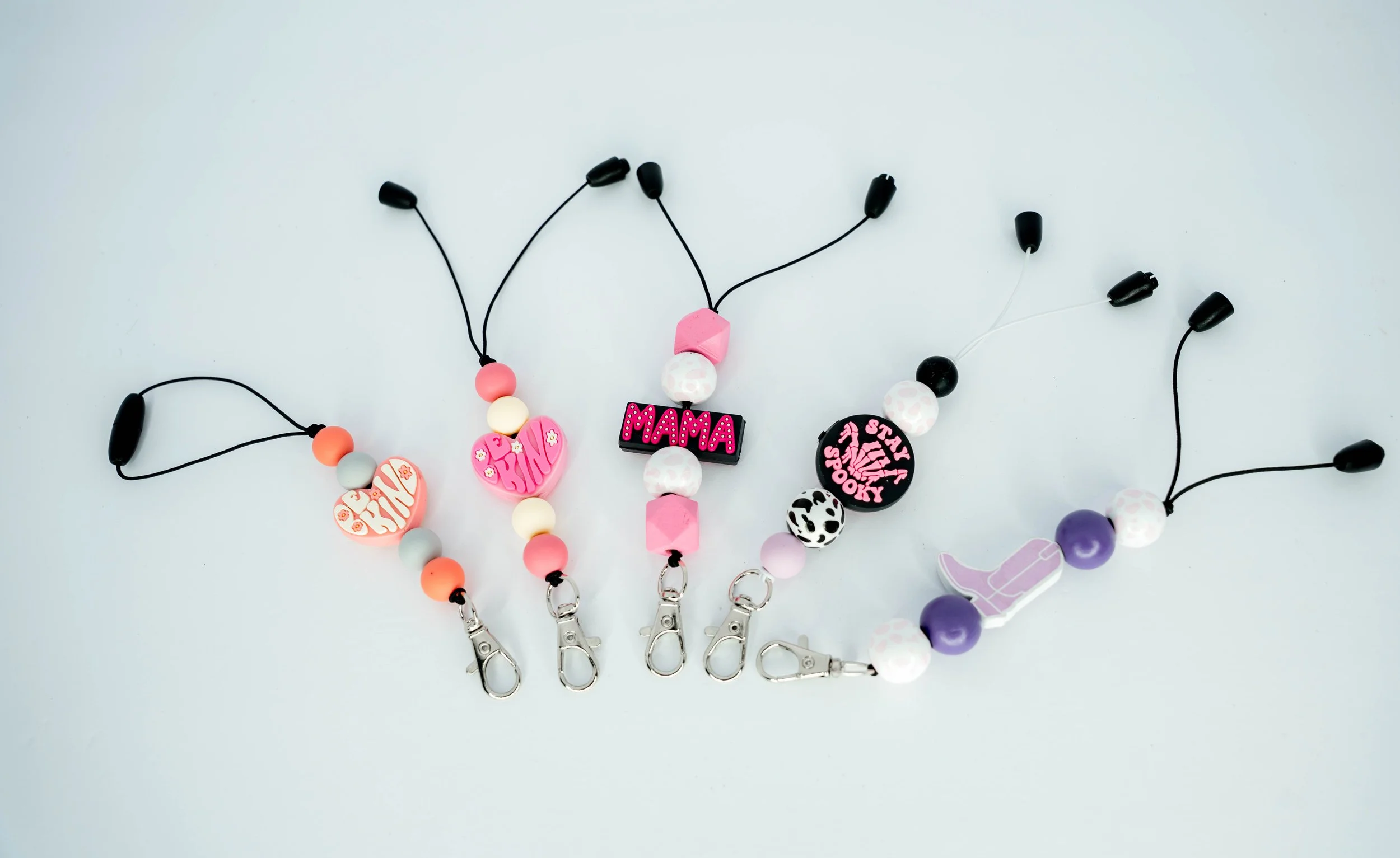 Five silicone teething toy pacifier clips with beads and charms, in pink, black, purple, and white, displayed on a light background.