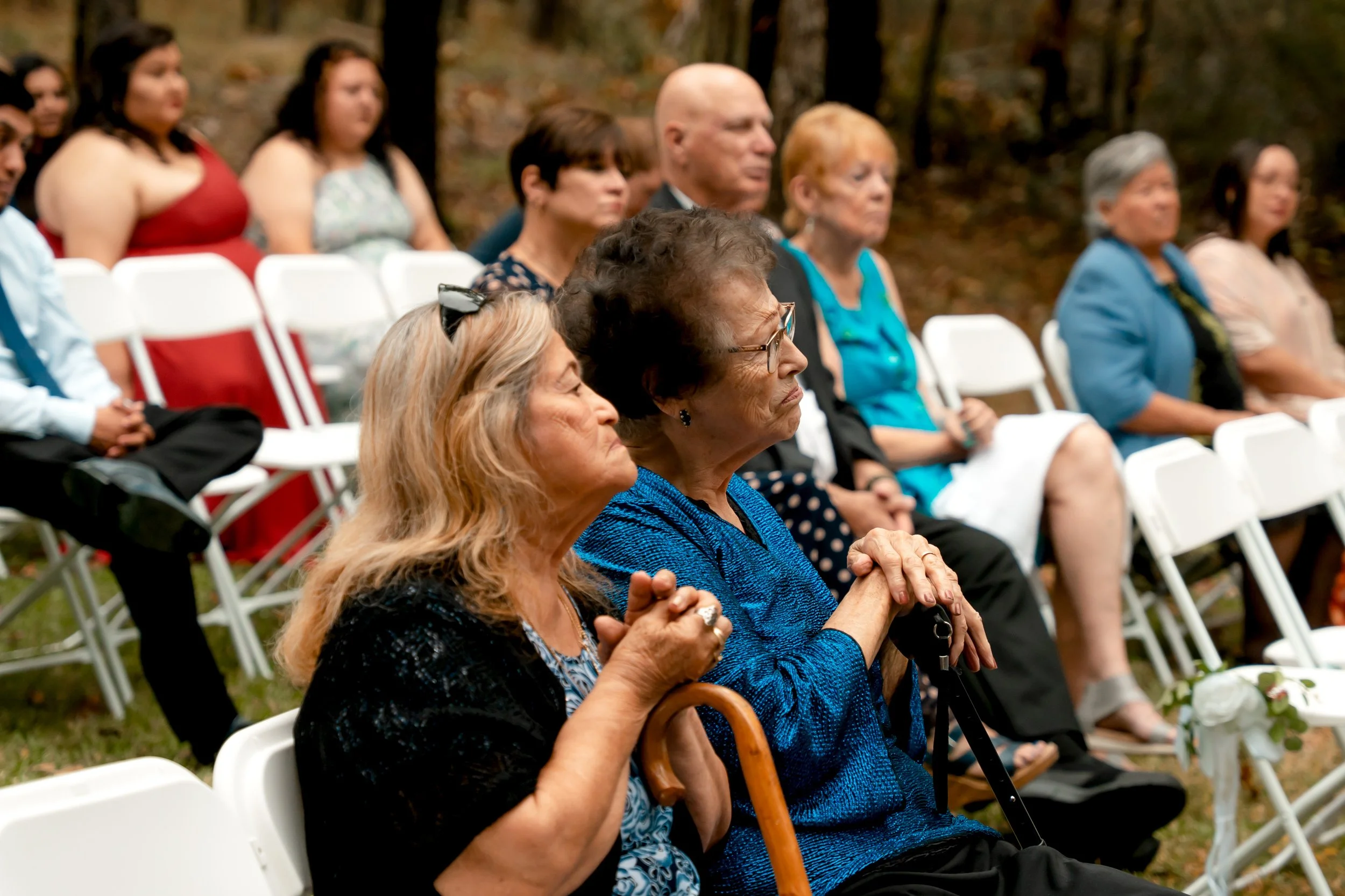Elderly woman with glasses and blue shirt and a woman with blond hair and a black cardigan sitting outdoors among other seated people at an event in a wooded area.