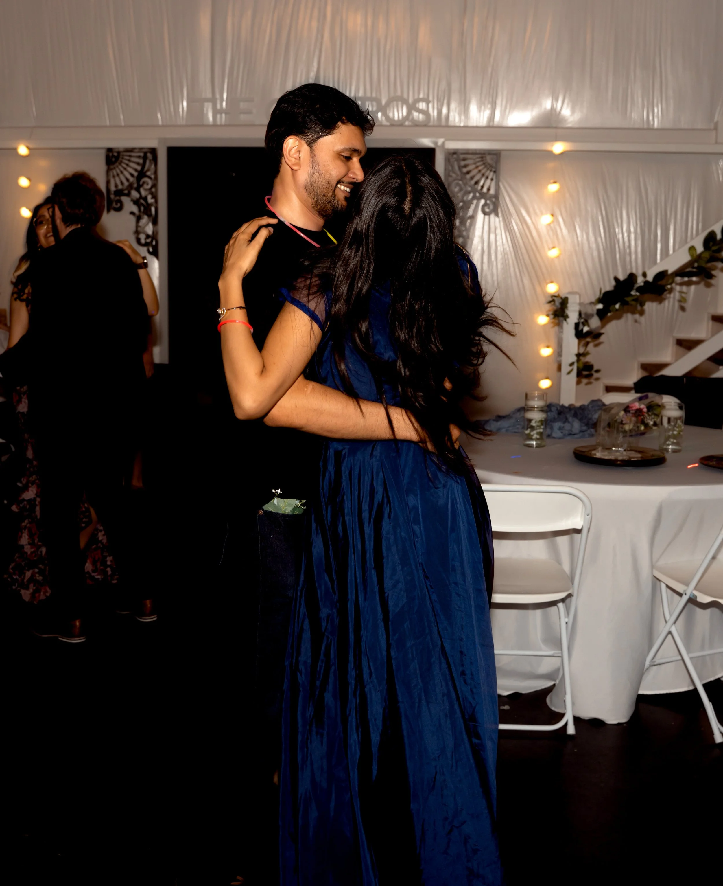A man and woman are dancing closely at an indoor event, with other people dancing in the background, in a decorated venue with string lights and a table with drinks.