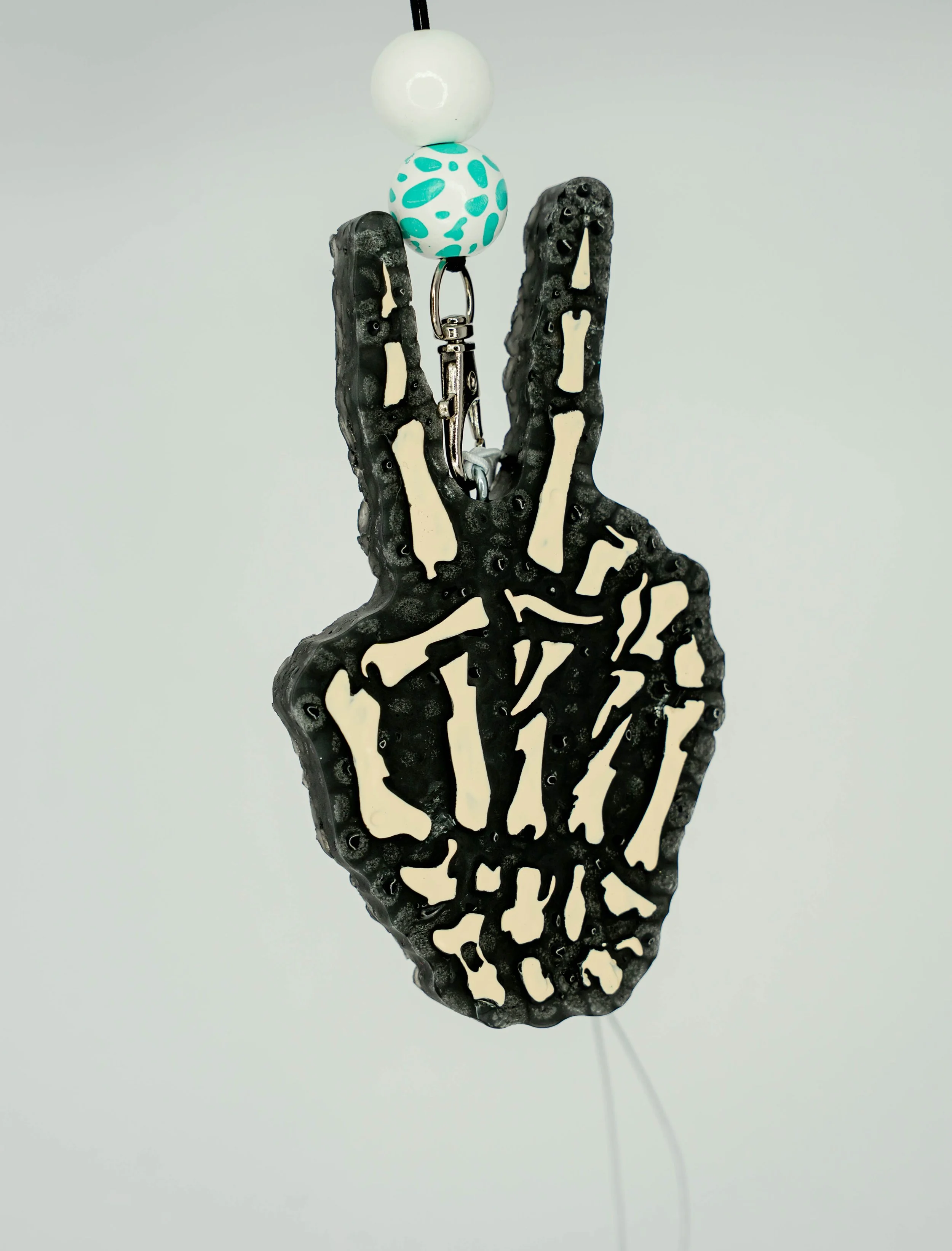 A black skeletal hand sculpture with white accents making a peace sign. It is hanging from a string with a small metal attachment, and above it are three decorative beads in white, teal, and white with teal spots.
