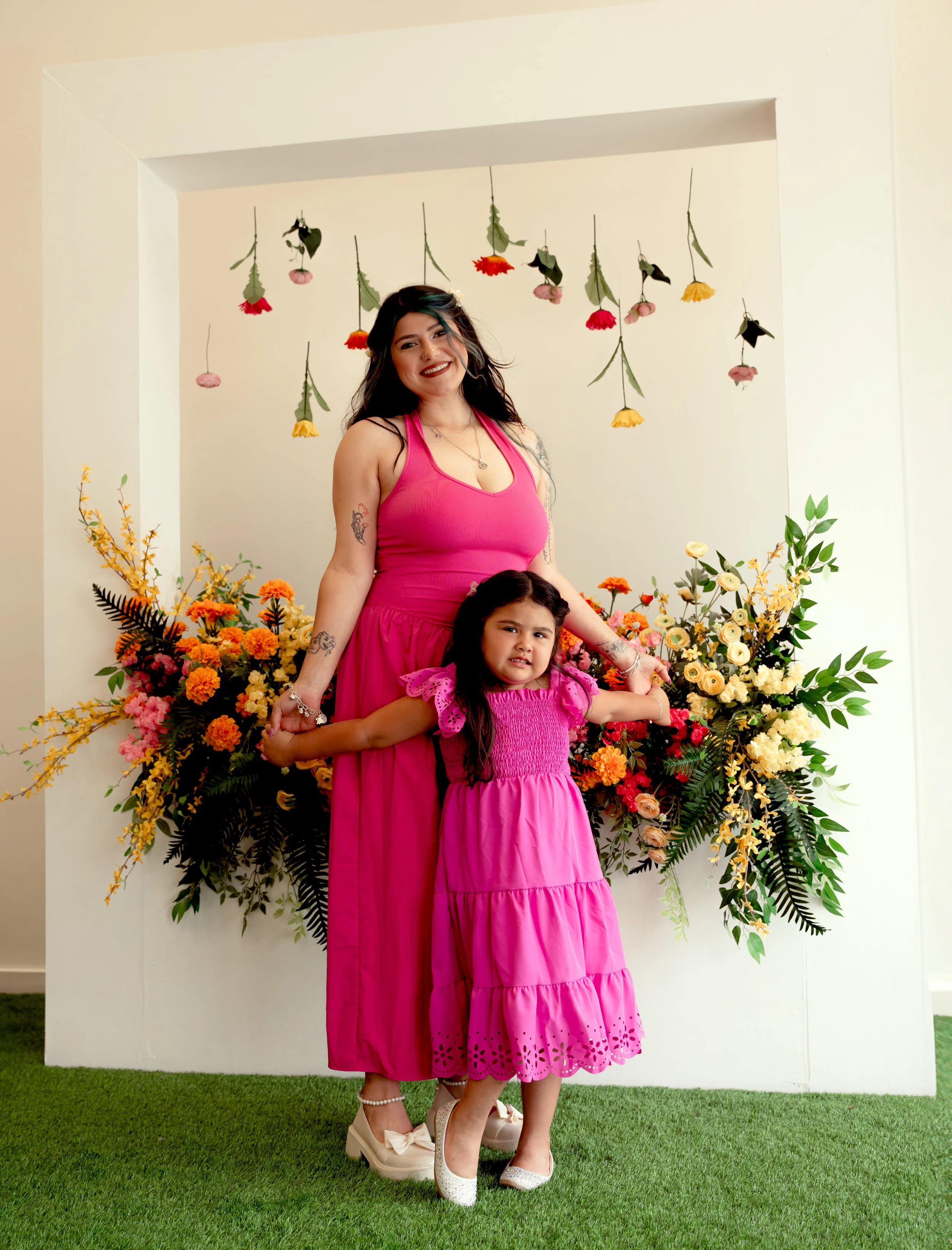 A woman and a young girl, both wearing pink dresses, stand together in front of a colorful floral arrangement and hanging flower decorations.