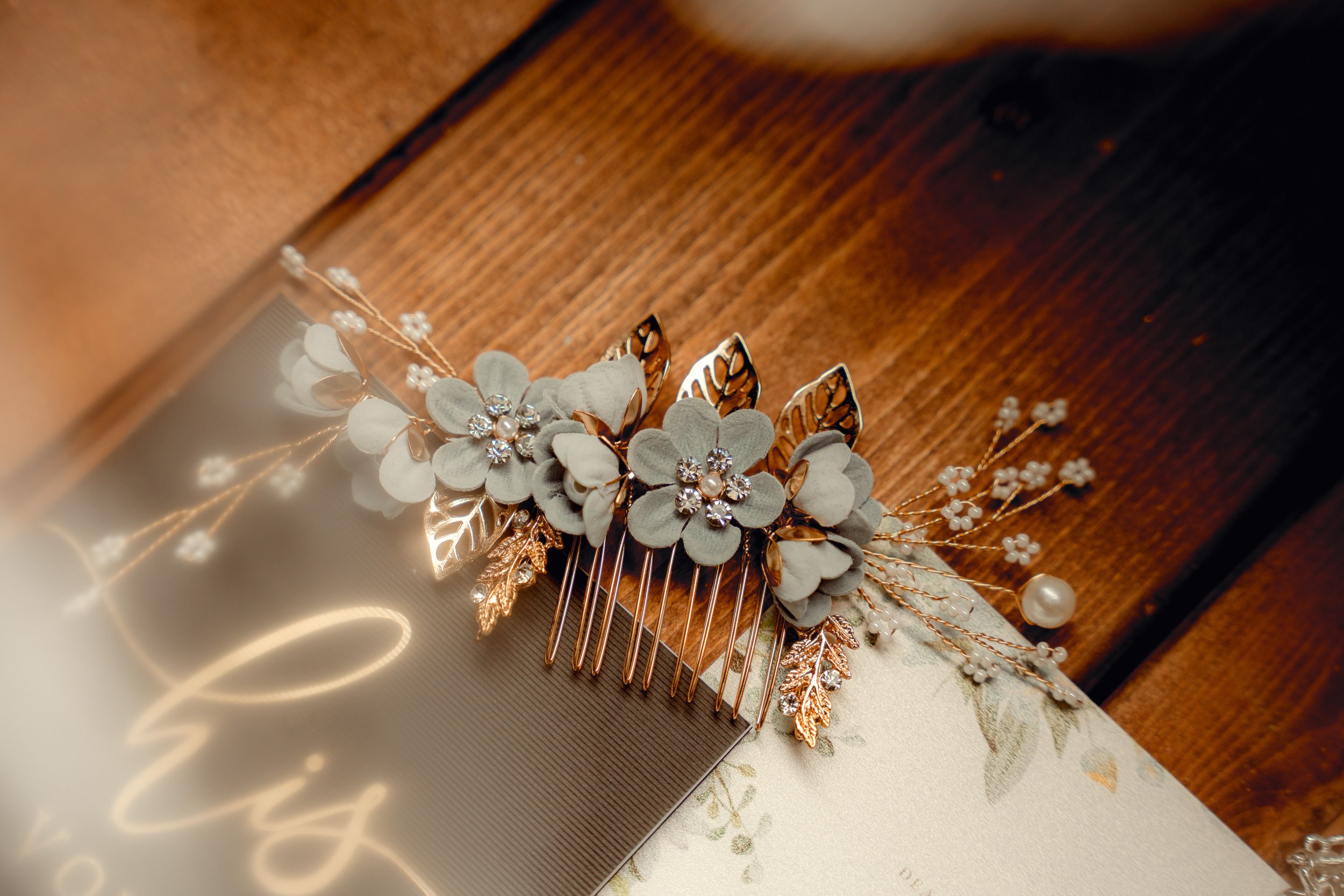 A decorative hair comb with white flowers, gold leaves, and pearl accents, placed on a wooden surface.