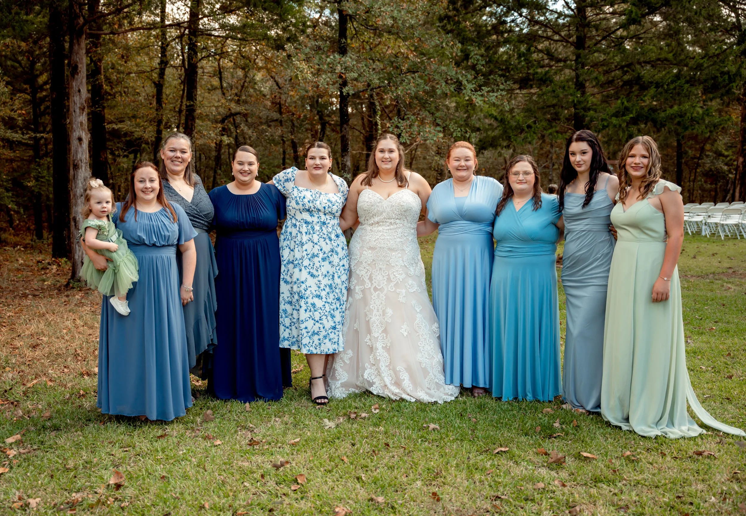 Group of women and a girl dressed in formal dresses standing outdoors in a wooded area with chairs in the background, celebrating a special occasion.