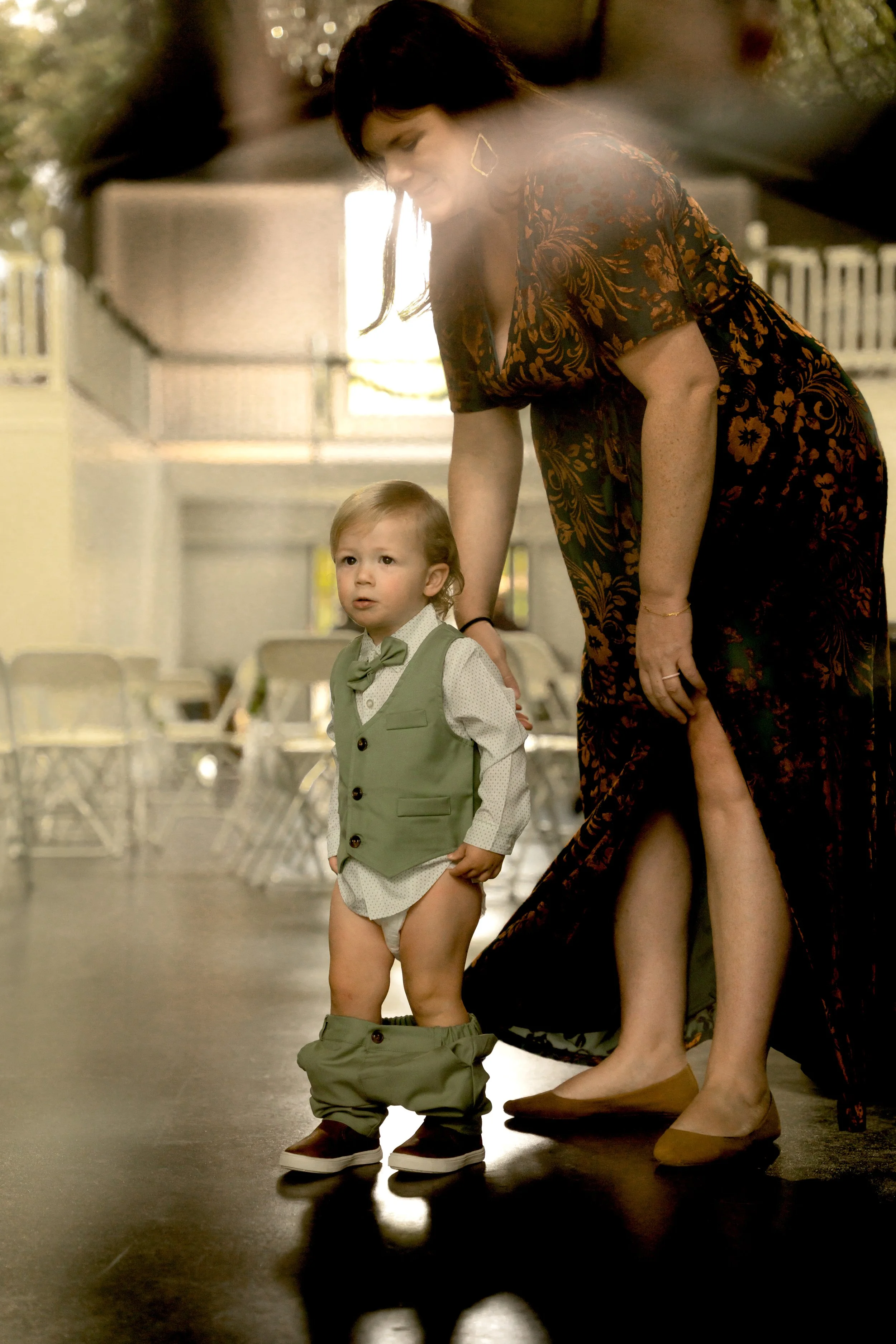 A woman helps a young boy put on his pants at an outdoor event, with chairs and a building in the background.
