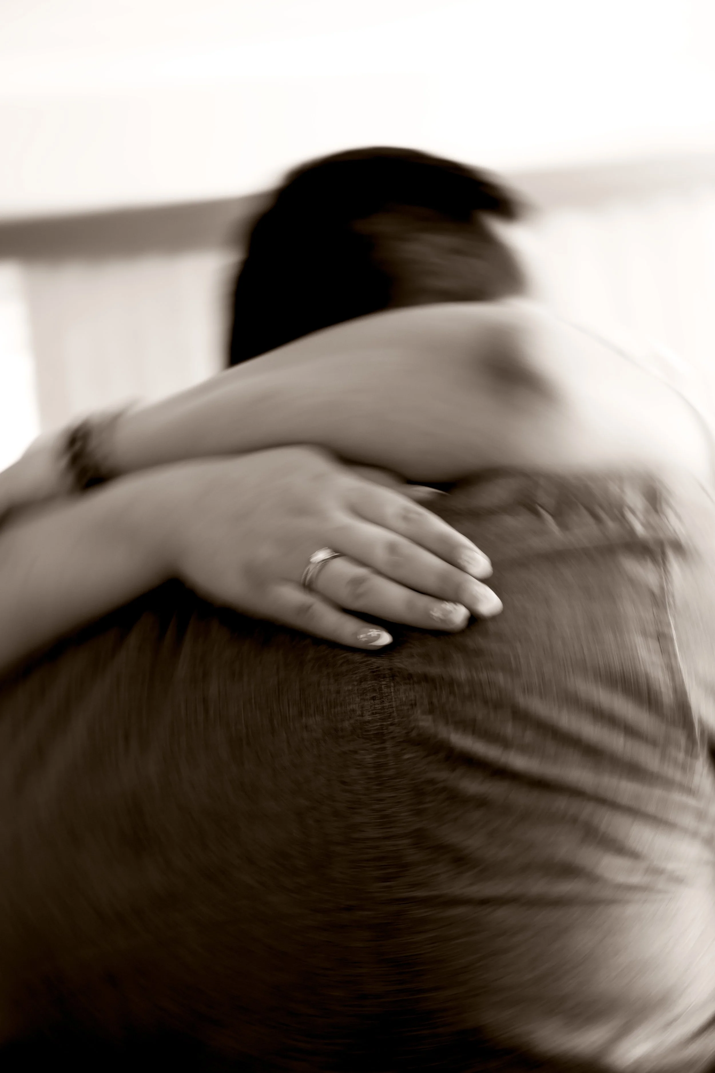 A couple embracing, with the woman hugging the man tightly. The photo is in black and white and slightly blurred, focusing on their embrace.