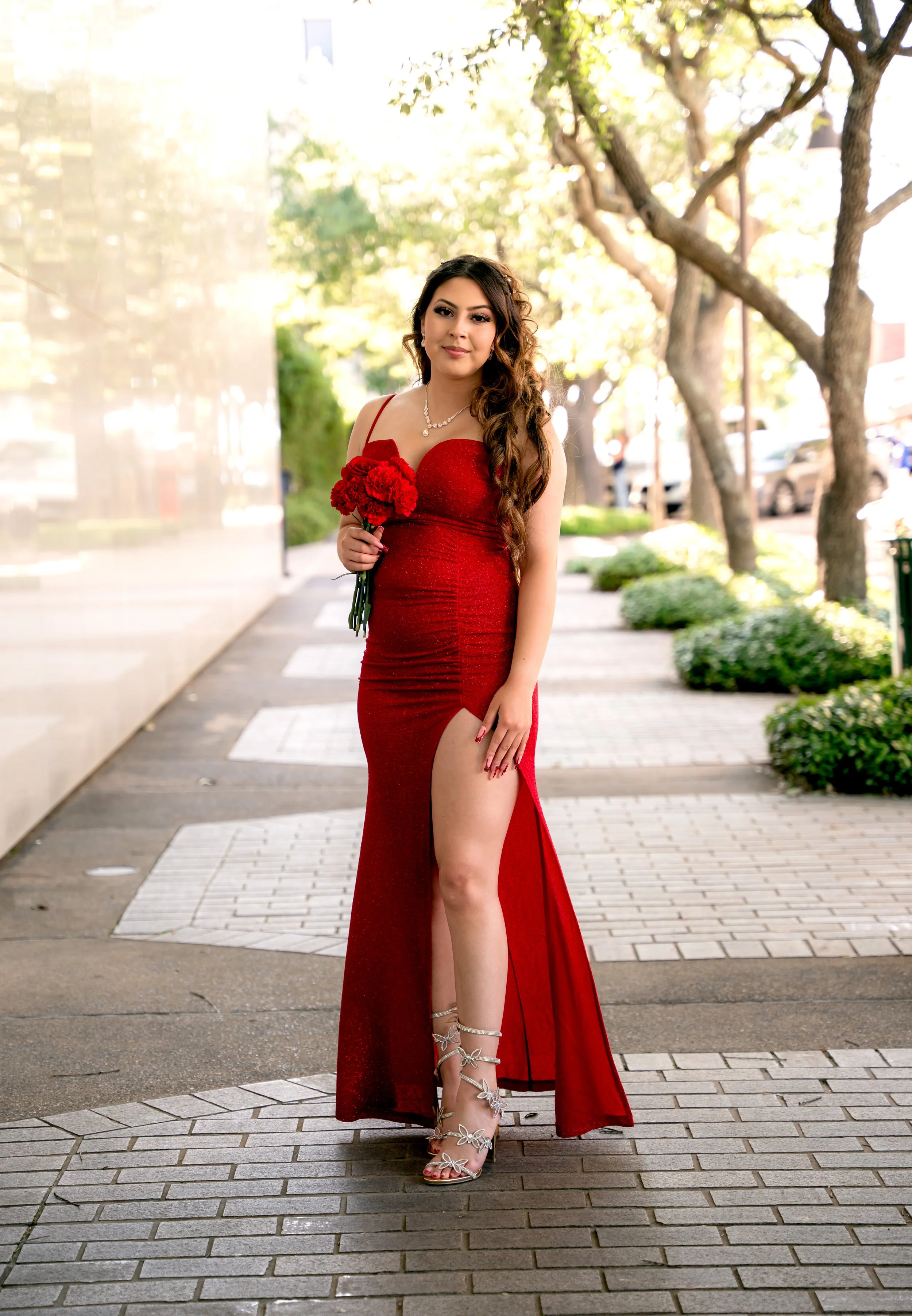 A young woman in a red dress holding a small bouquet of red flowers, standing on a sidewalk with trees and parked cars in the background, during daylight.