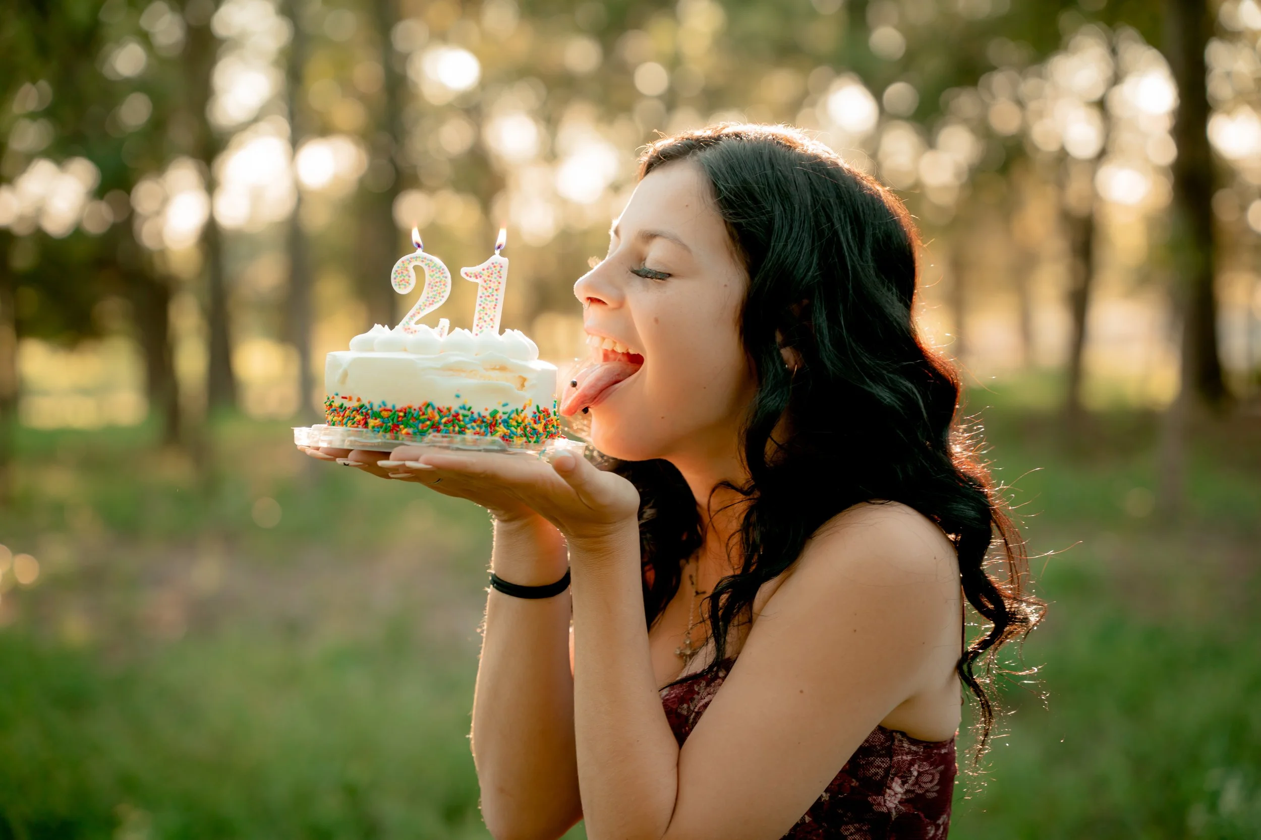 A young woman with black wavy hair is holding a birthday cake with lit candles shaped as 21 outdoors in a park during sunset. She is licking the cake, celebrating her 21st birthday.