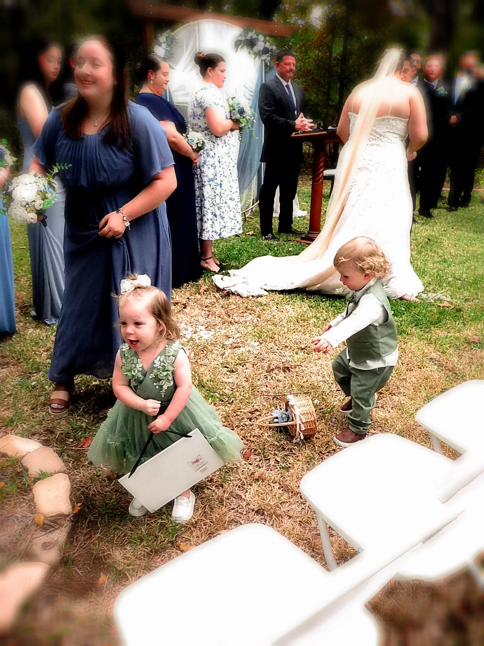 Children and adults gathered outdoors at a wedding ceremony, with a bride in a white gown and veil, and a woman in a blue dress holding a bouquet. Two young children are in foreground, one girl in a green dress and boy in a vest and pants, playing on