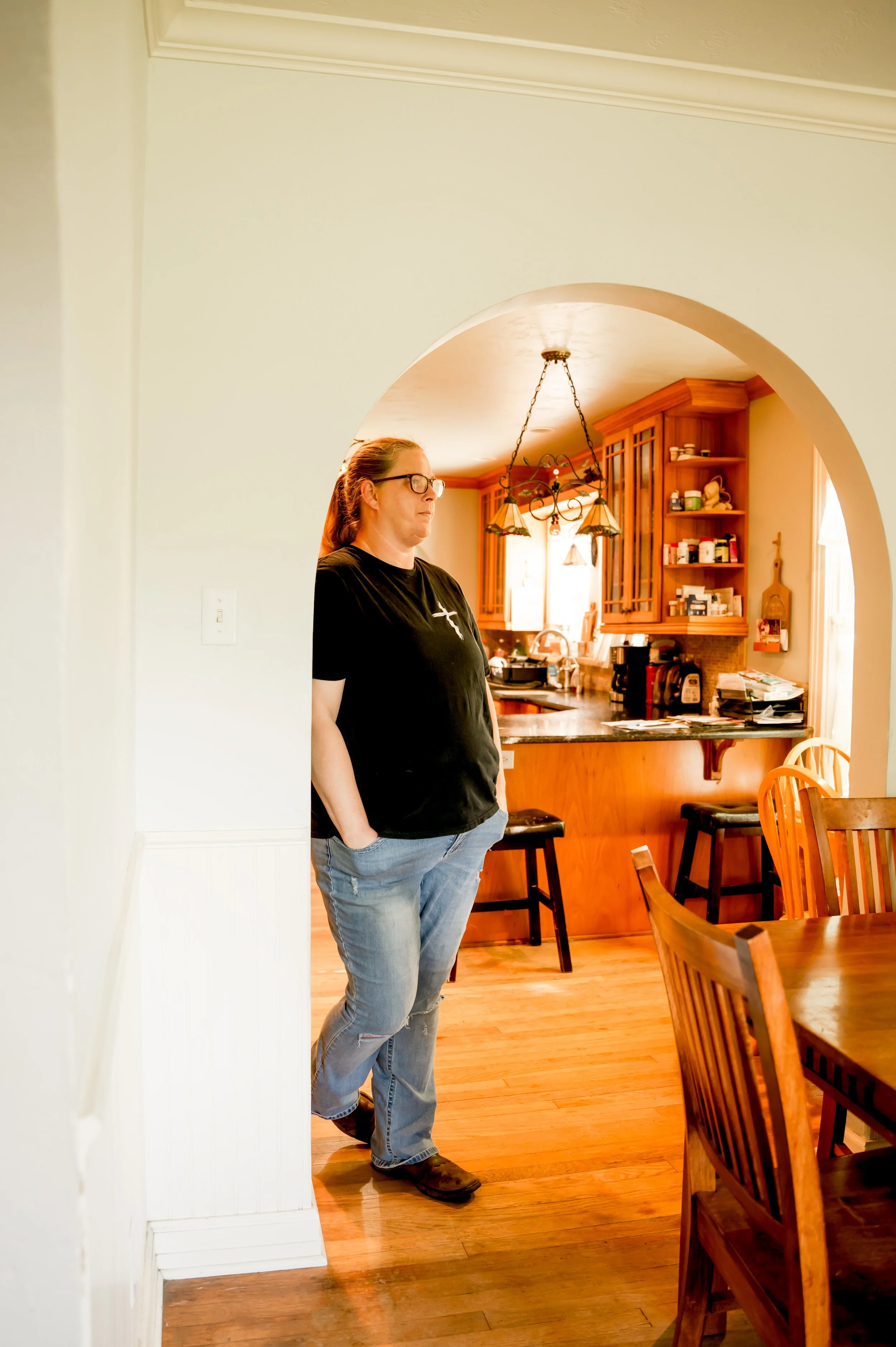 A woman with glasses and long hair, wearing a black t-shirt and jeans, standing in a kitchen area, leaning against a wall with her hands in her pockets.