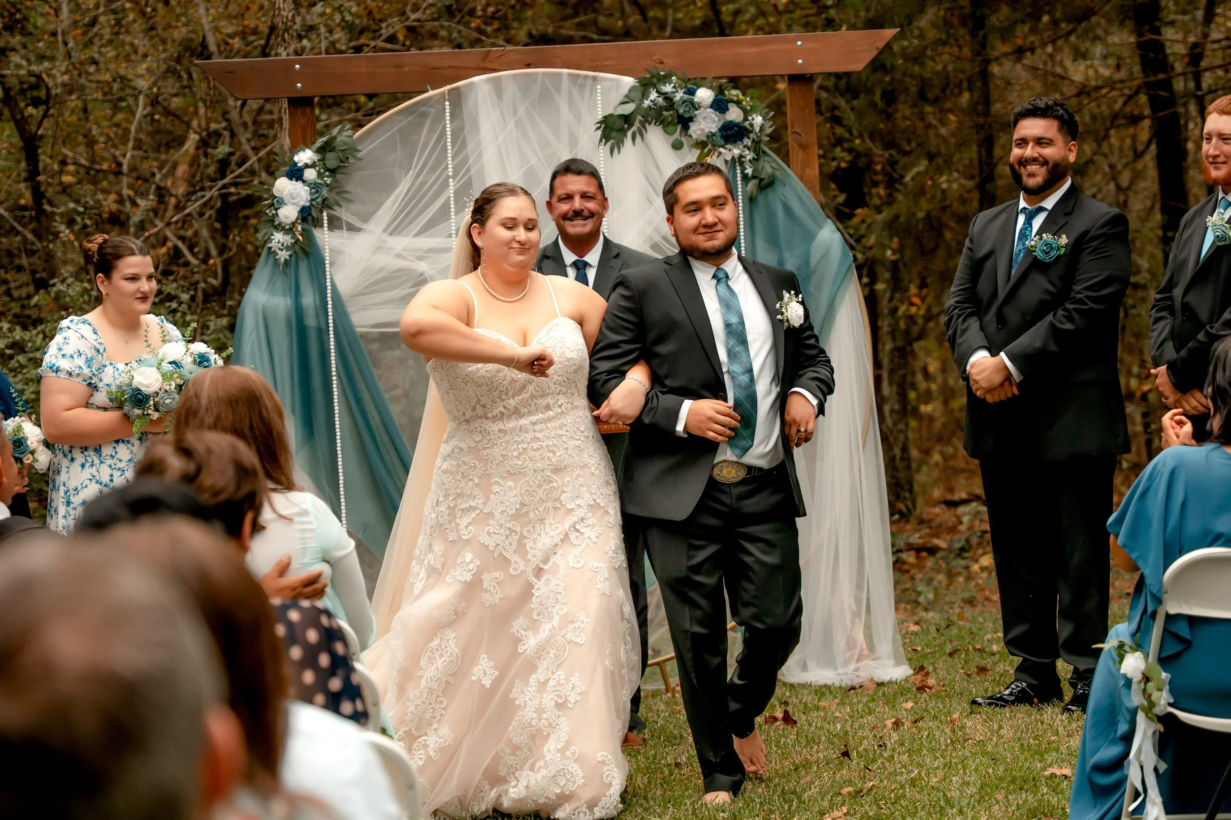 A wedding ceremony outdoors with the bride and groom walking arm in arm past the officiant, surrounded by bridesmaids and groomsmen, with guests seated watching. The scene features a decorated arch and autumn trees in the background.