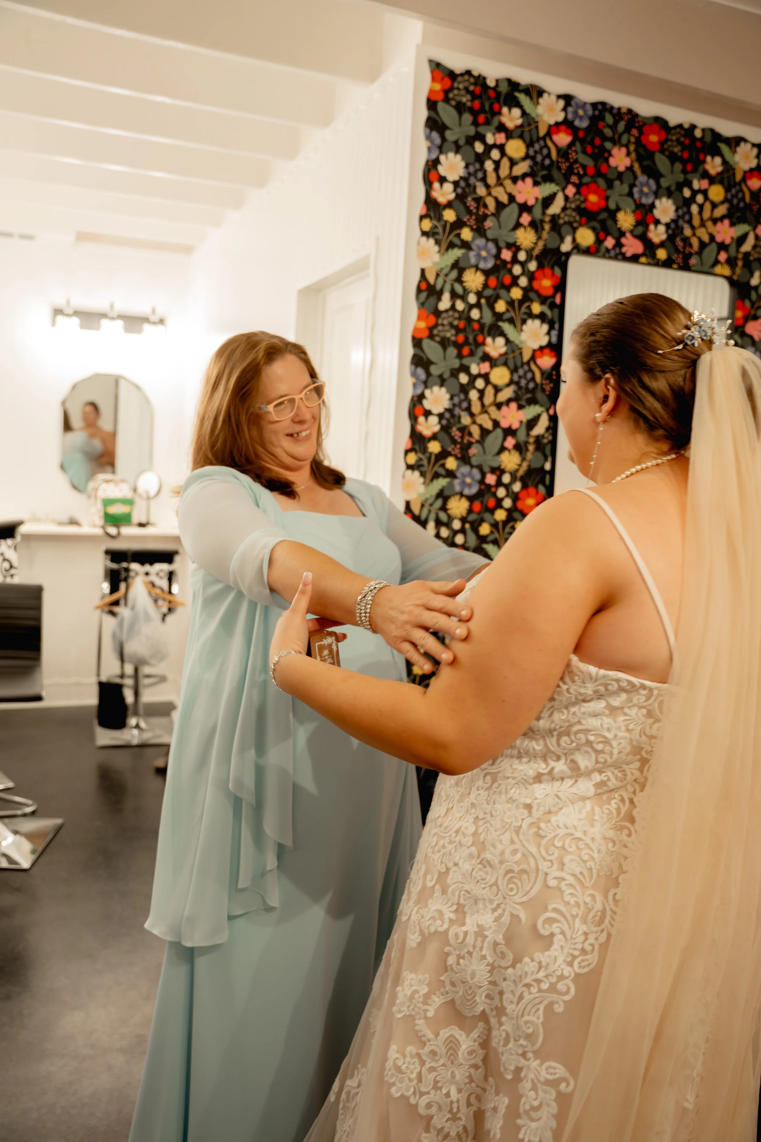 A bride in a lace wedding dress and veil smiling as she holds hands with a woman in a pale blue dress, inside a room with floral artwork on the wall.