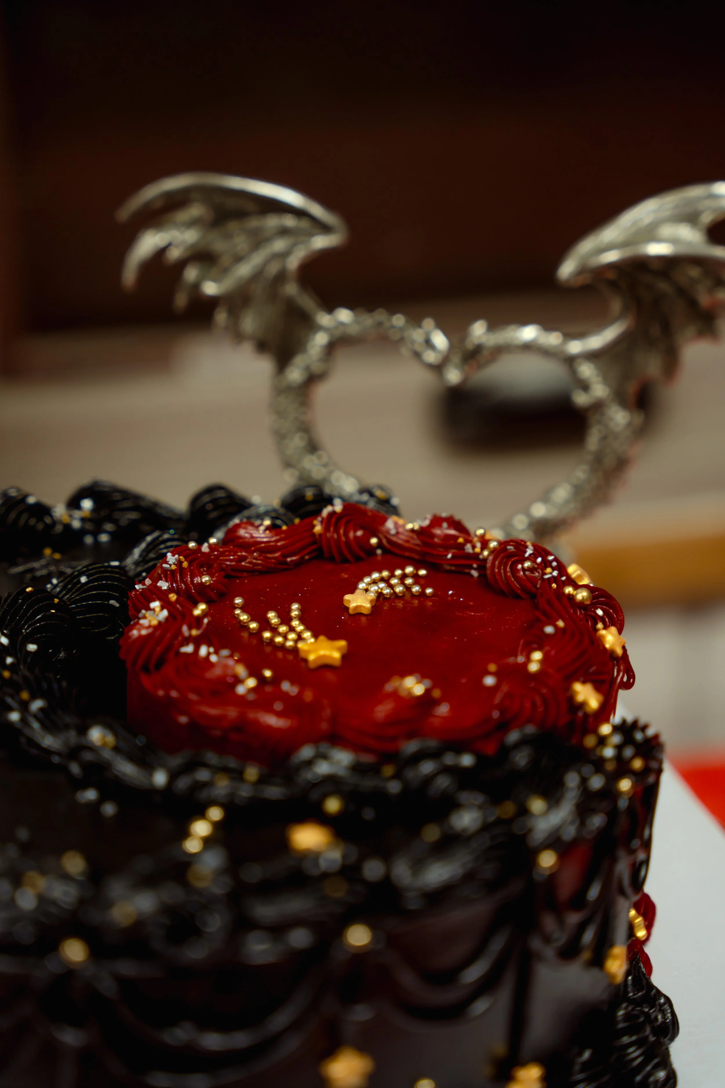 A decorated black and red cake with gold and white sprinkles, with a metallic dragon heart-shaped decoration with wings in the background.