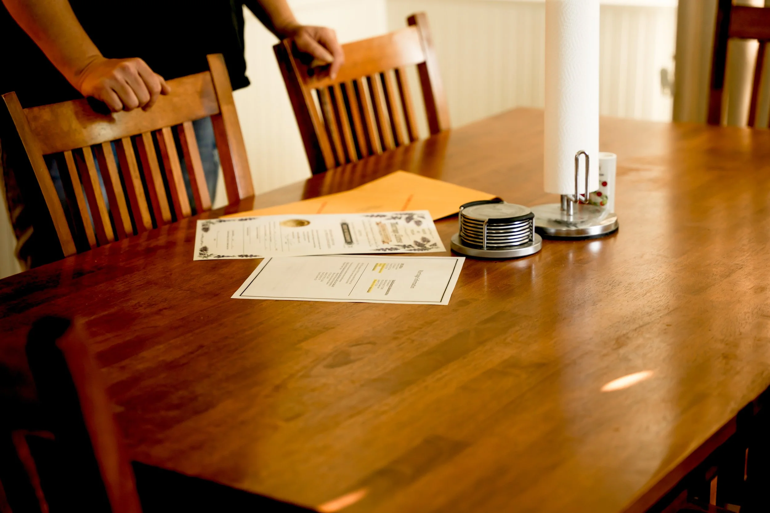 A wooden dining table with menus, a paper towel holder, and a condiment container, with person's hands resting on the back of a chair.