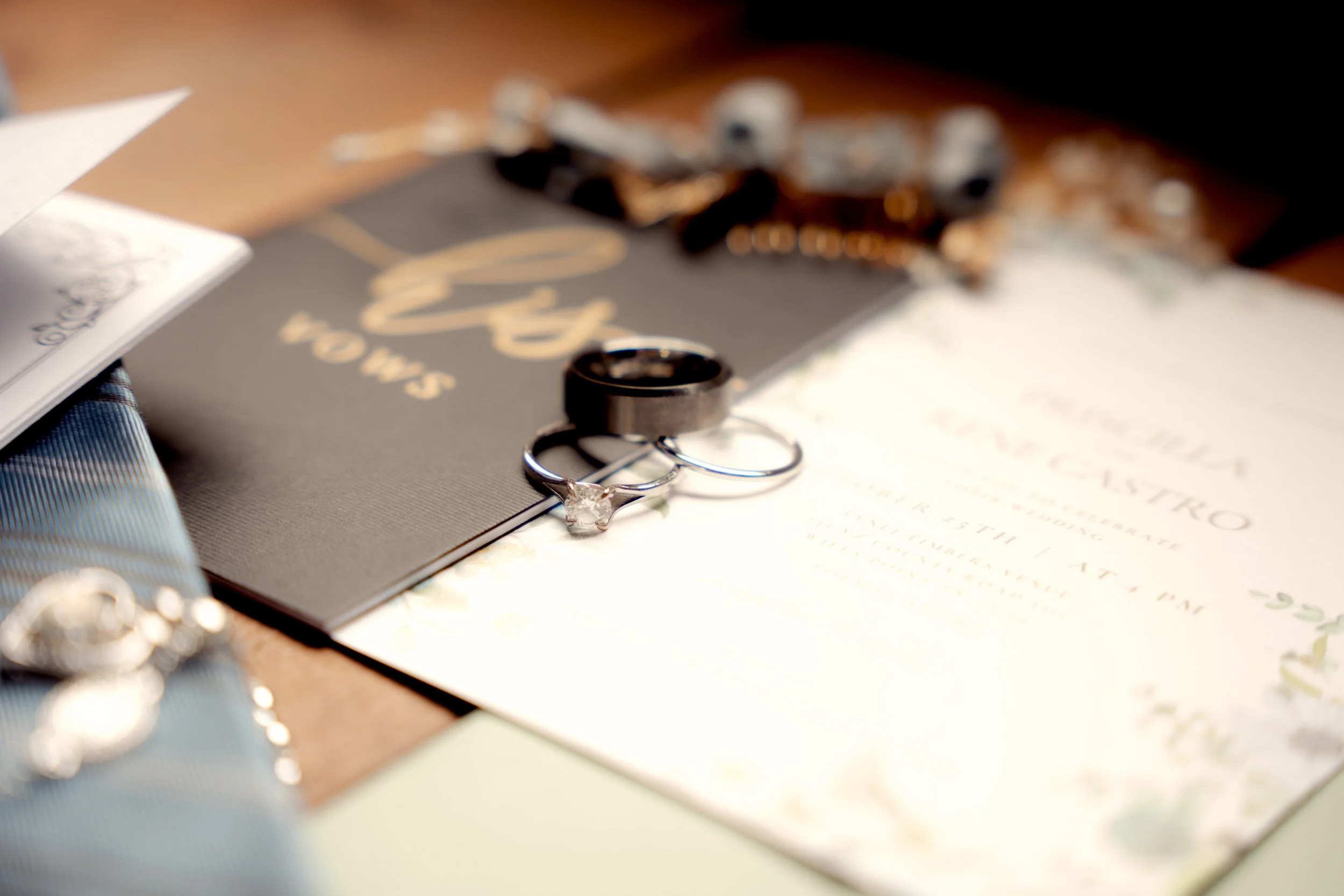 Close-up of wedding rings, including one with a diamond, placed on a wedding invitation, with a black card and jewelry in the blurred background.