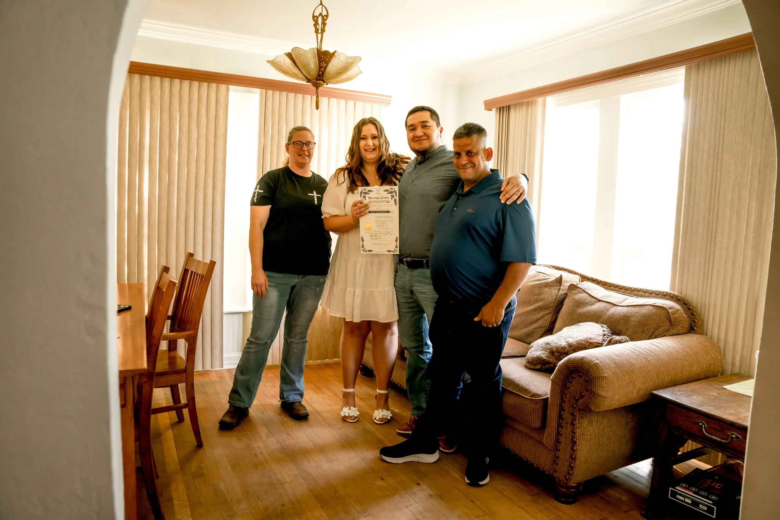 Group of four people smiling and posing inside a living room, with one woman holding a certificate, standing in front of a beige sofa with cushions. The room has wood flooring, large windows with vertical blinds, and a chandelier hanging from the cei