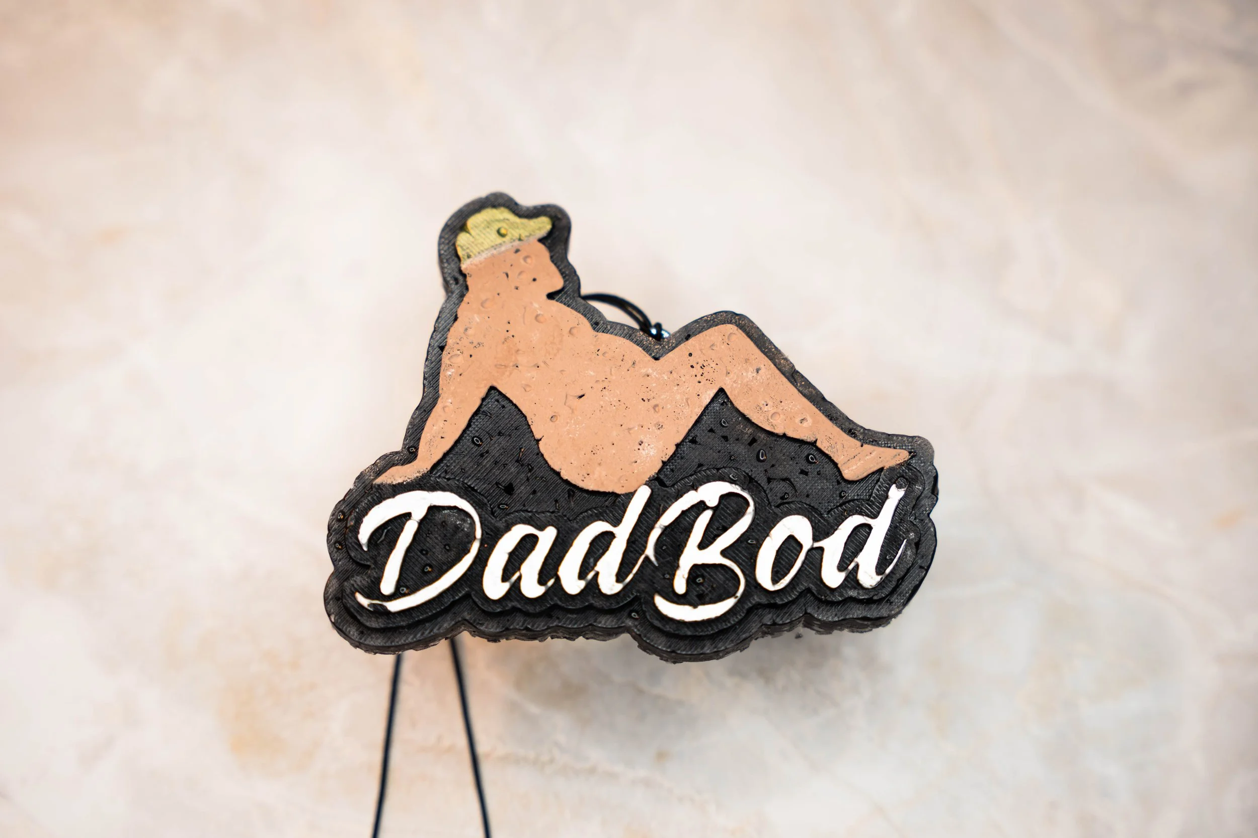 A decorative patch with a cartoon-style reclining nude man wearing a hat, above the word "Dadbod" in script font.