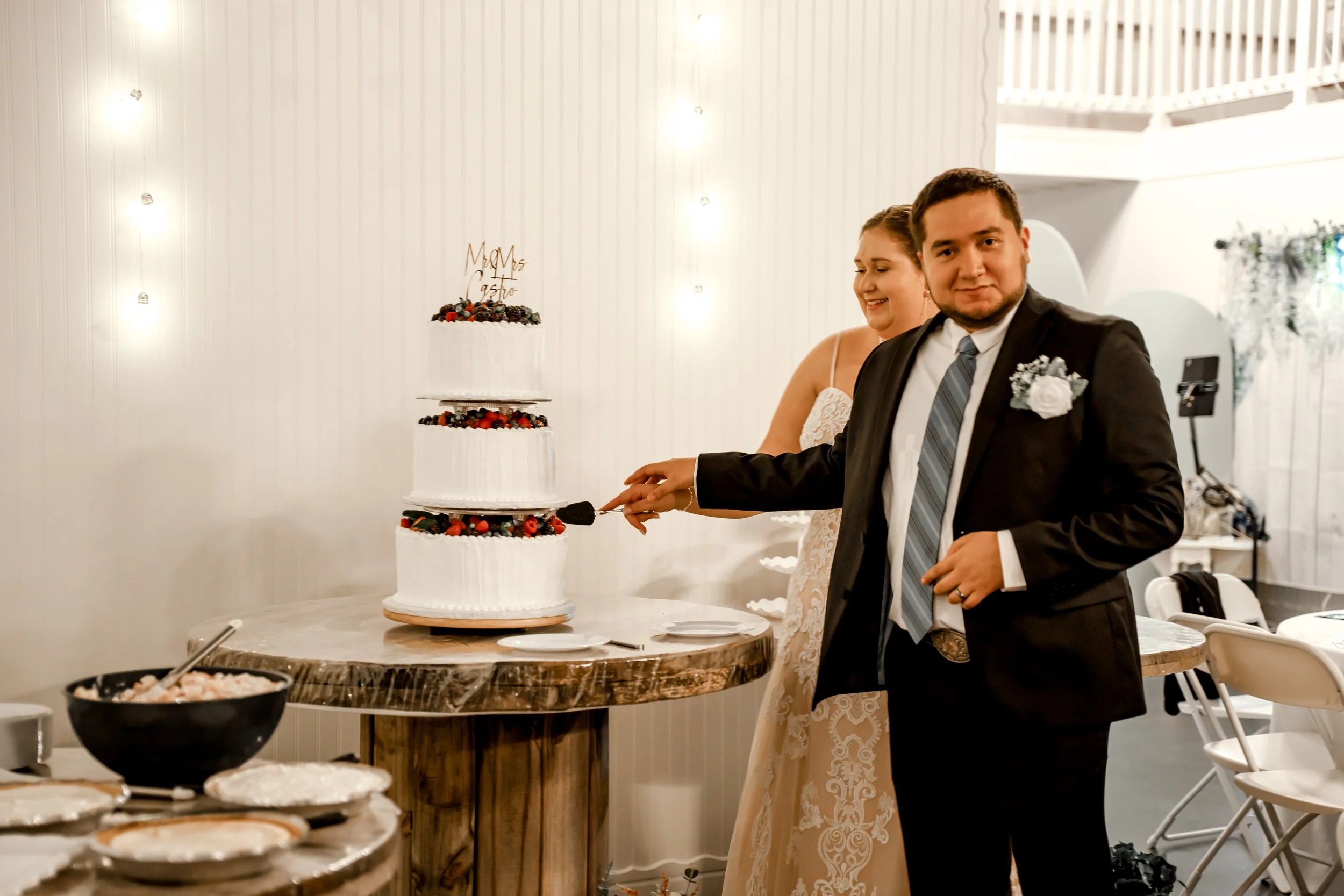 A man in a suit slices a wedding cake with a woman in a white dress standing behind him at a wedding reception.