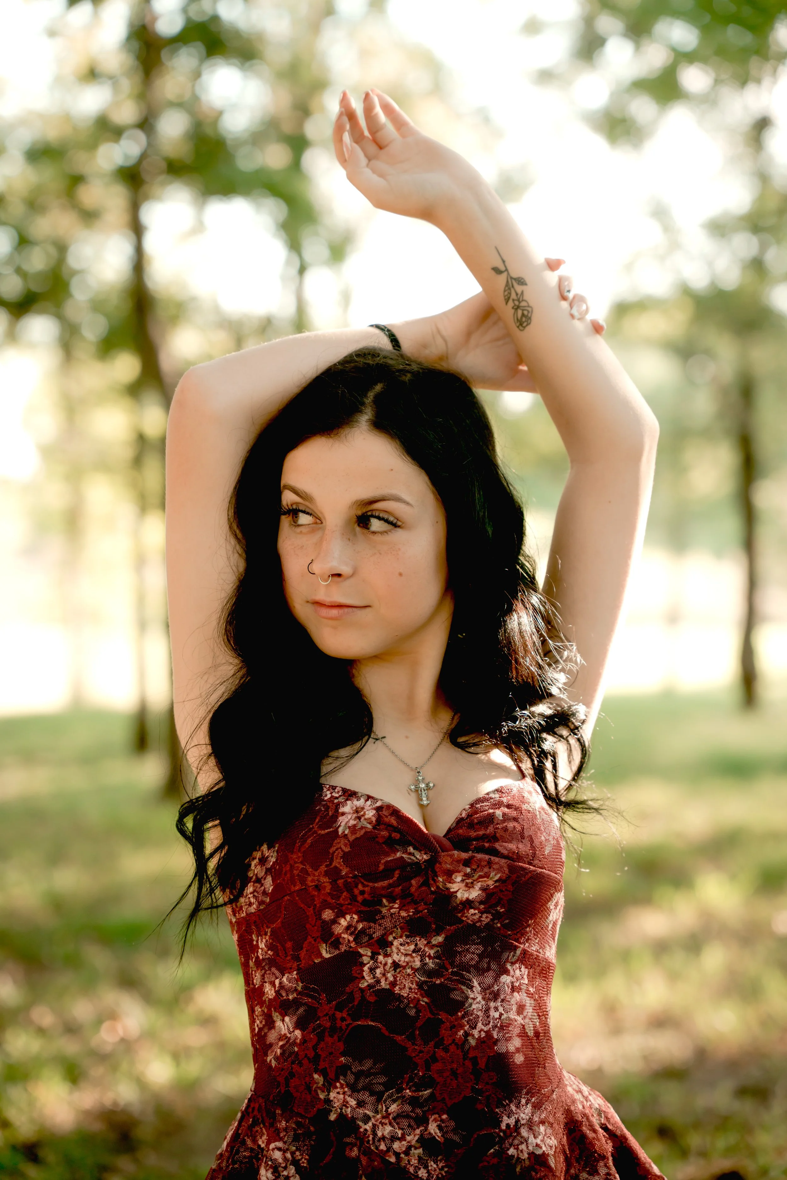 A young woman with black hair and tattoos raises her arms above her head, standing outdoors in a wooded area on a sunny day.