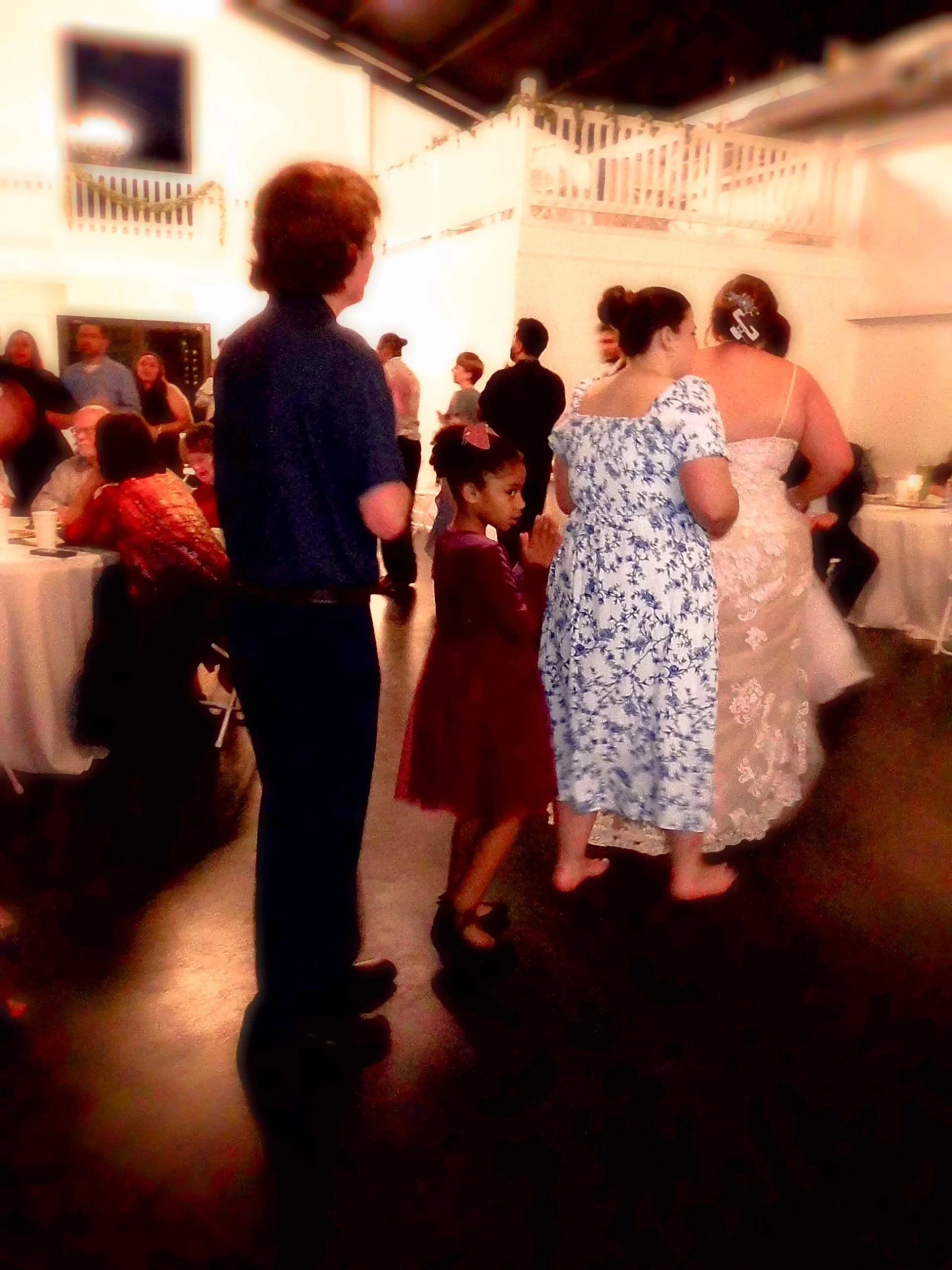 People at a wedding reception, including a young girl in a maroon dress, a woman in a floral dress, and a bride in a white wedding gown, in an indoor event space with tables and guests in the background.