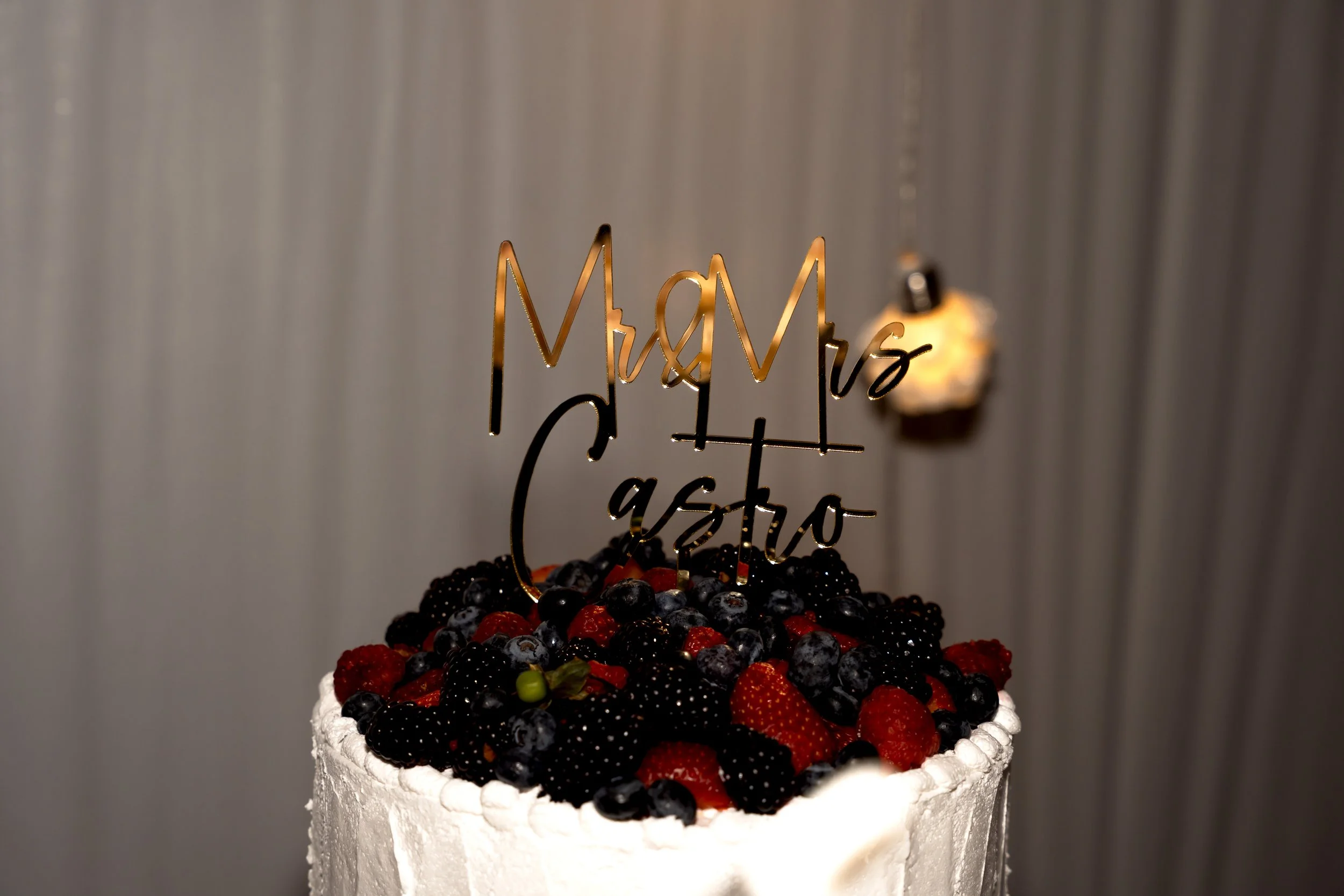 A wedding cake topped with assorted berries and a gold cake topper that reads 'Mr & Mrs vs Caspo' against a textured silver background.
