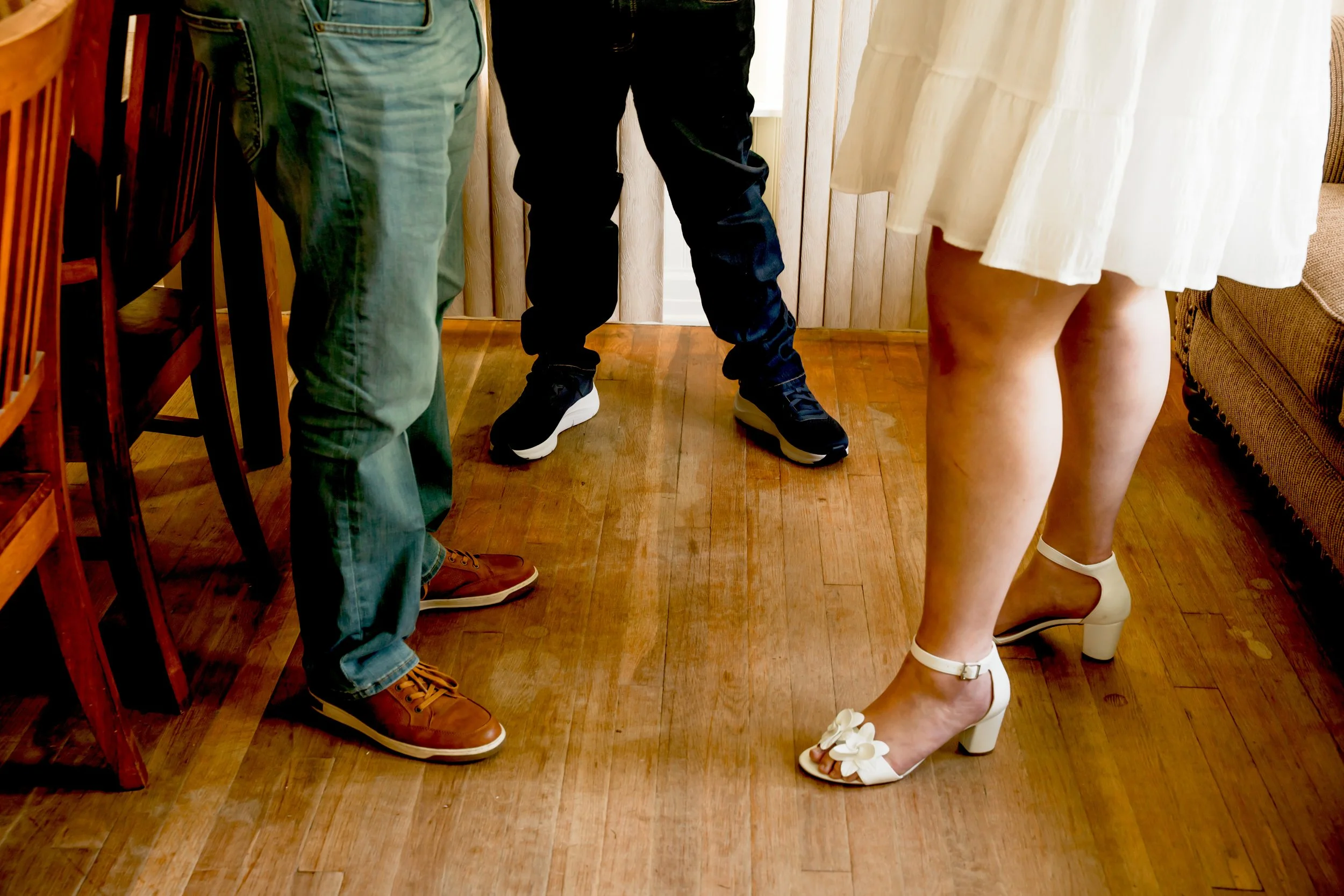 Close-up of three people standing on a wooden floor, showing their lower bodies and feet. The person on the left is wearing jeans and brown sneakers, the person in the middle is in black pants and black sneakers, and the person on the right is wearin