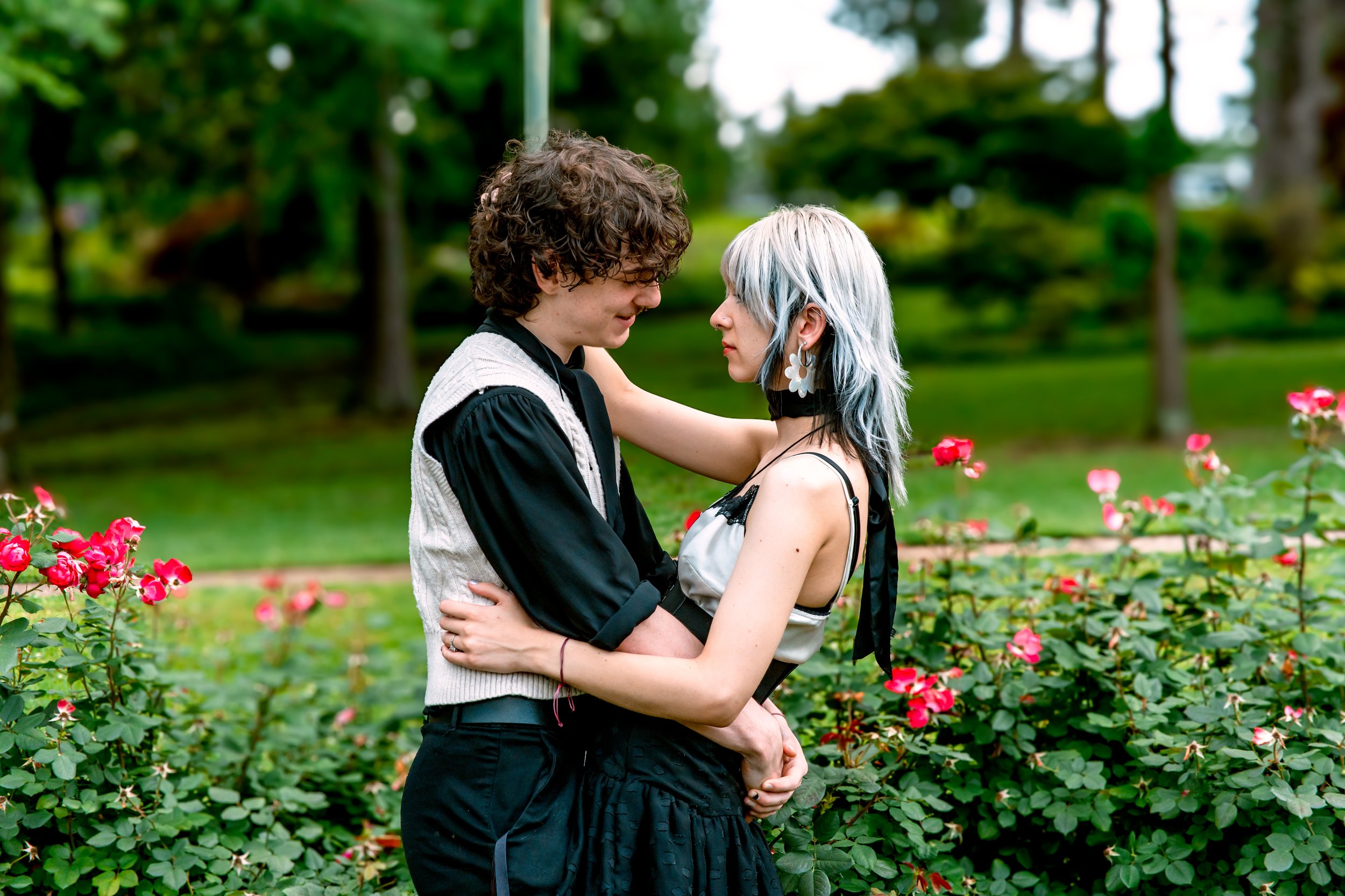 A young man and woman embrace in a park with pink roses and green foliage.