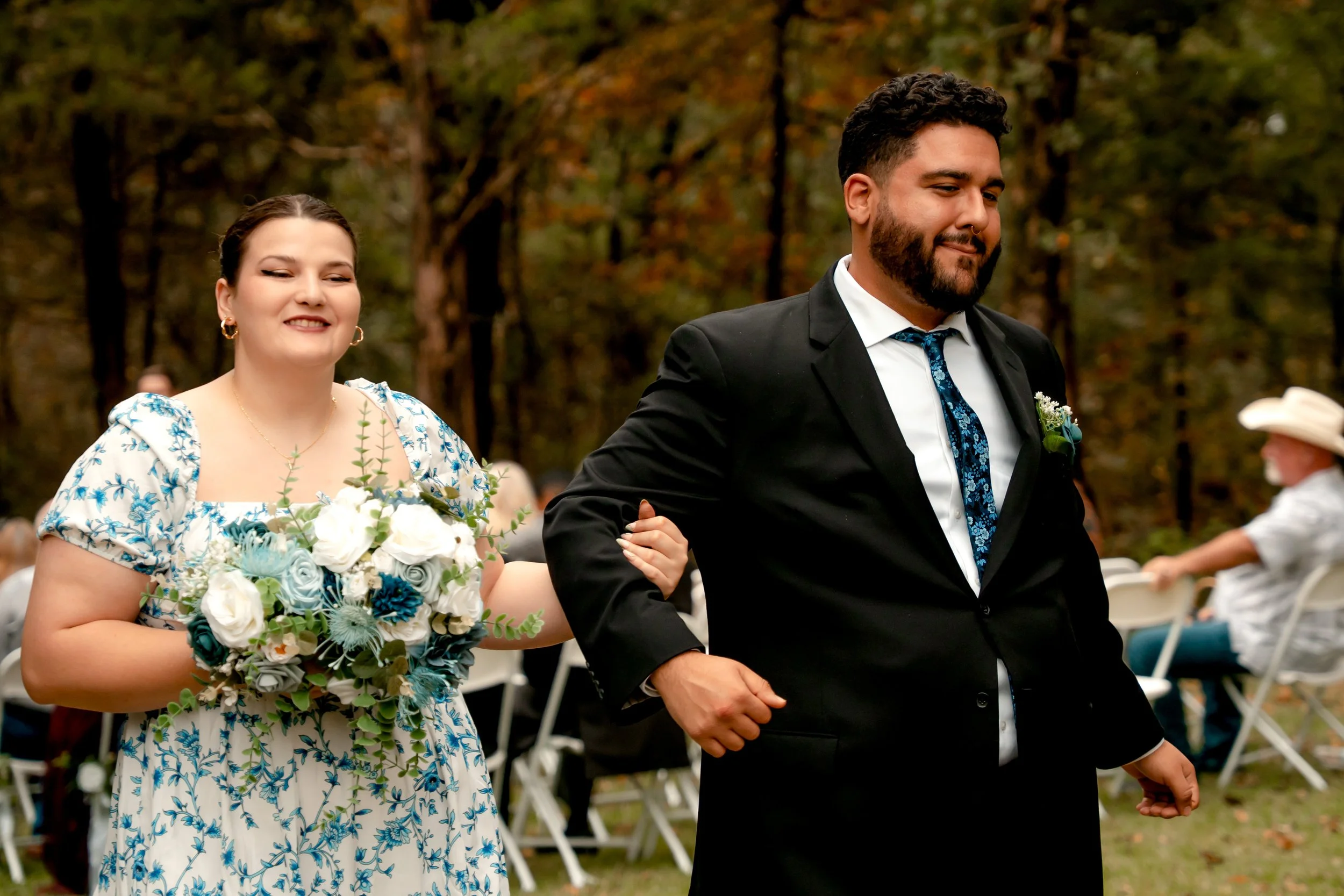 A newlywed couple walking outdoors at a wedding ceremony in a wooded area. The bride is wearing a blue and white floral dress and holding a bouquet, and the groom is in a black suit with a blue tie.