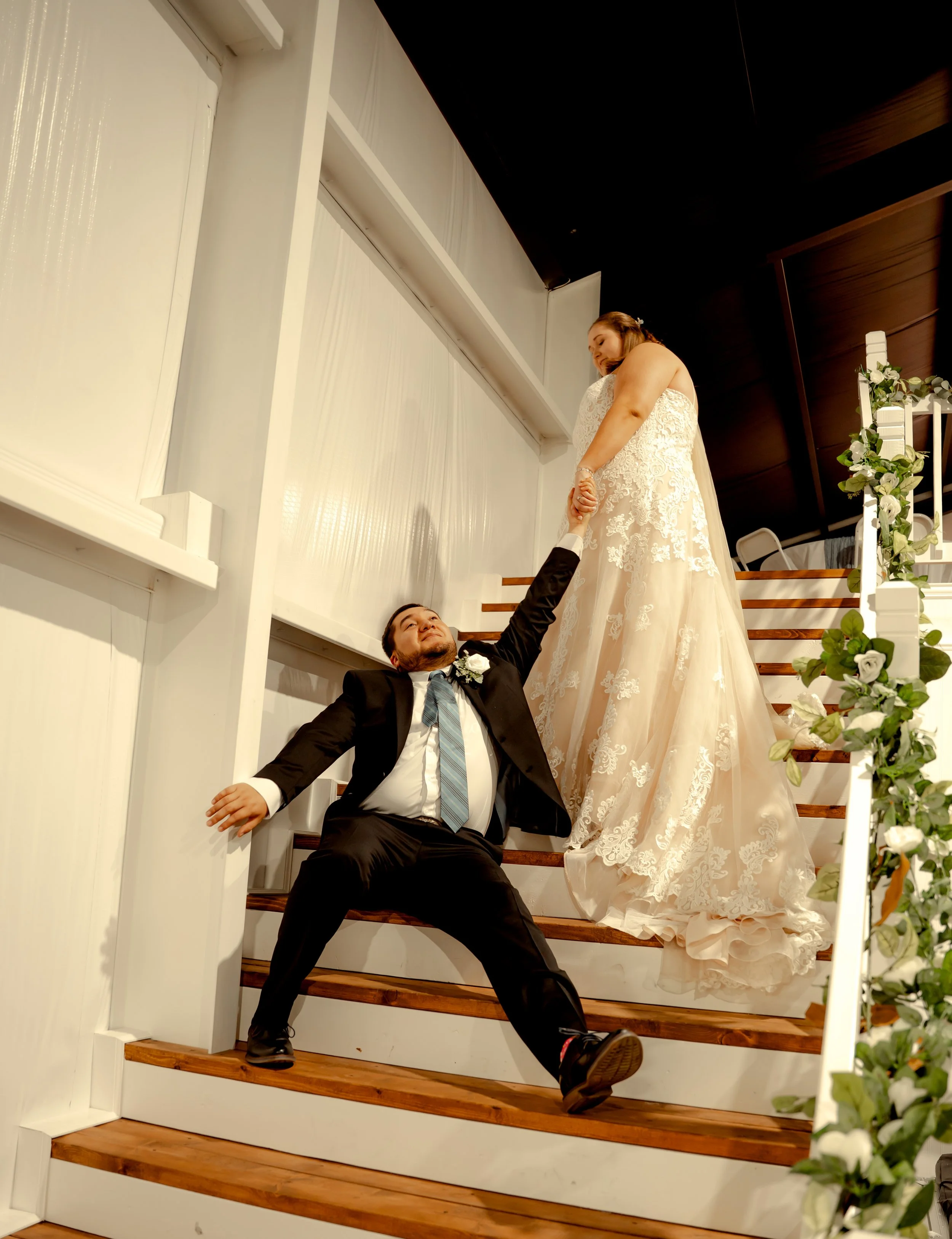 A bride and groom on a staircase, with the groom sitting on the stairs and reaching up to hold the bride's hand while she stands above him.