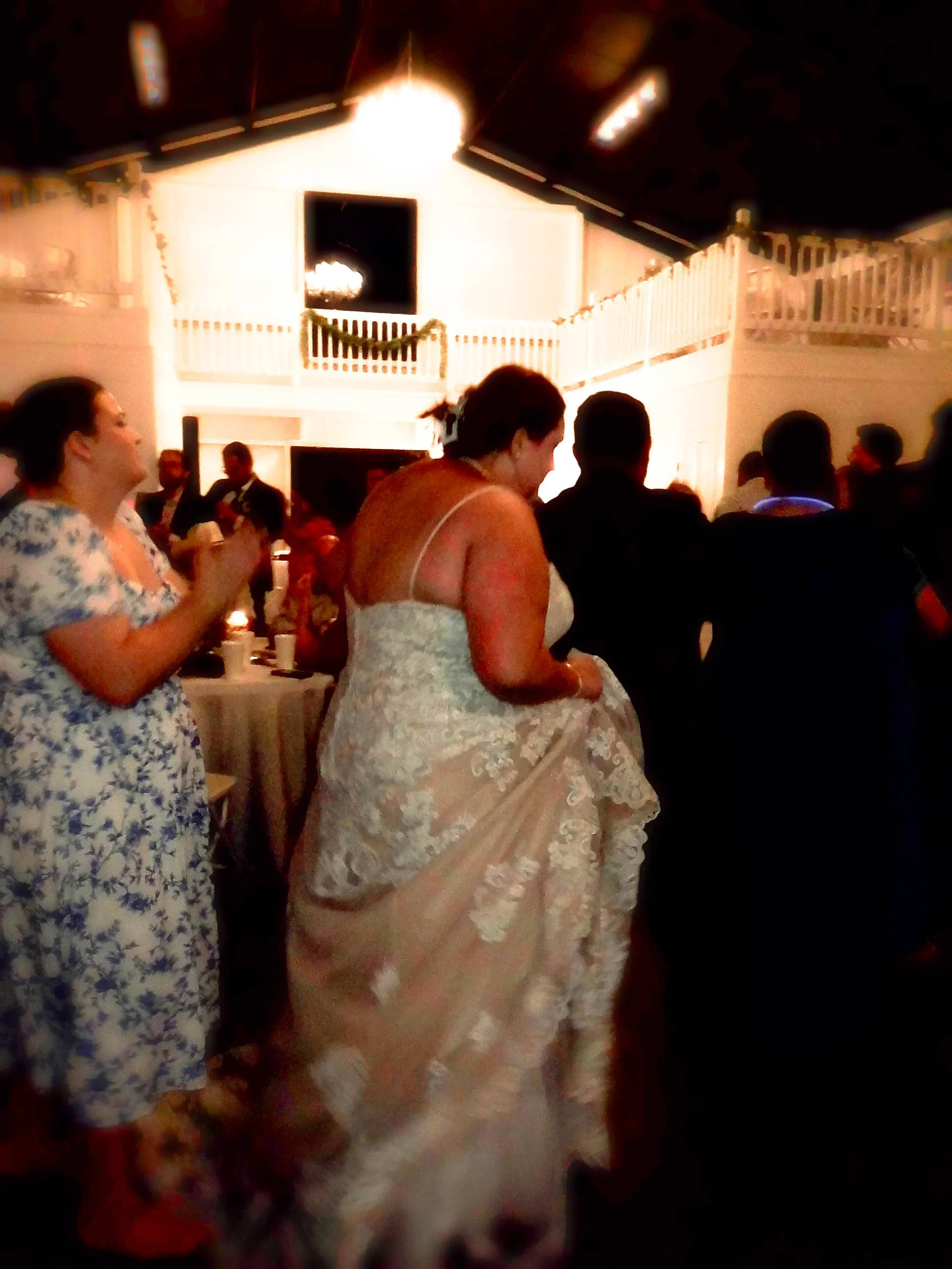 A crowded indoor wedding reception with guests dancing and celebrating. A woman in a white lace wedding dress is lifting her skirt in the center. The atmosphere is lively with dim lighting and some candles on tables.