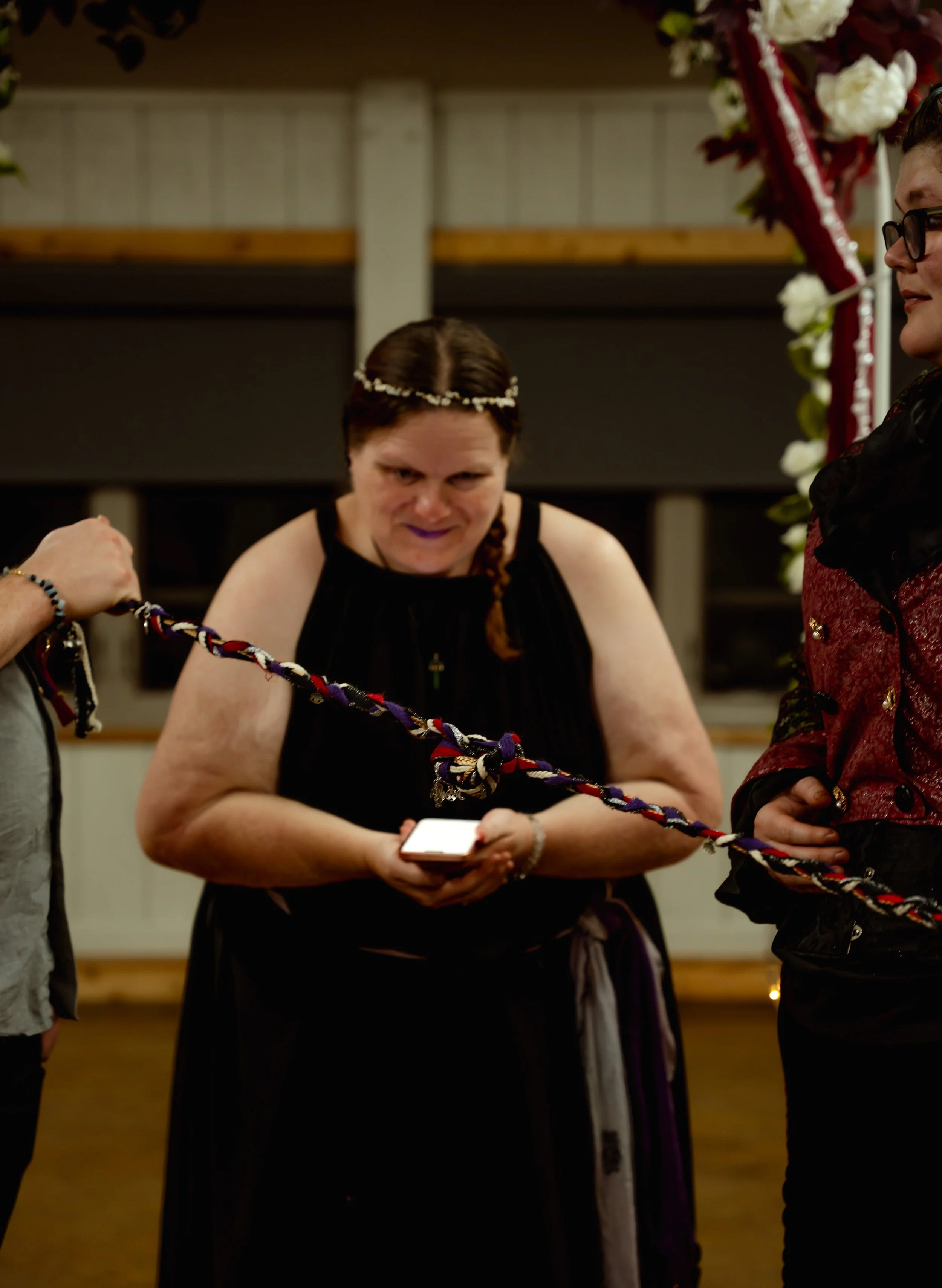 Woman in black dress looking at her phone holding a rope at a formal event, decorated with flowers and ribbon.