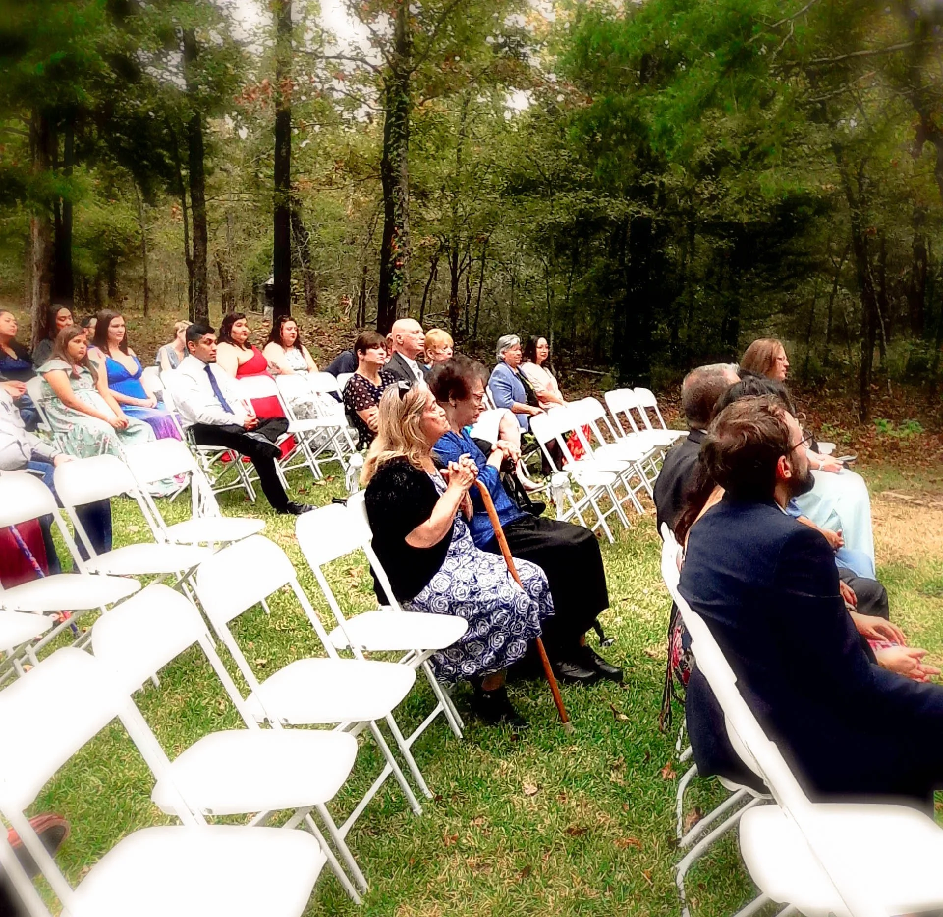 People seated outdoors on white folding chairs in a wooded area, attending a formal event.