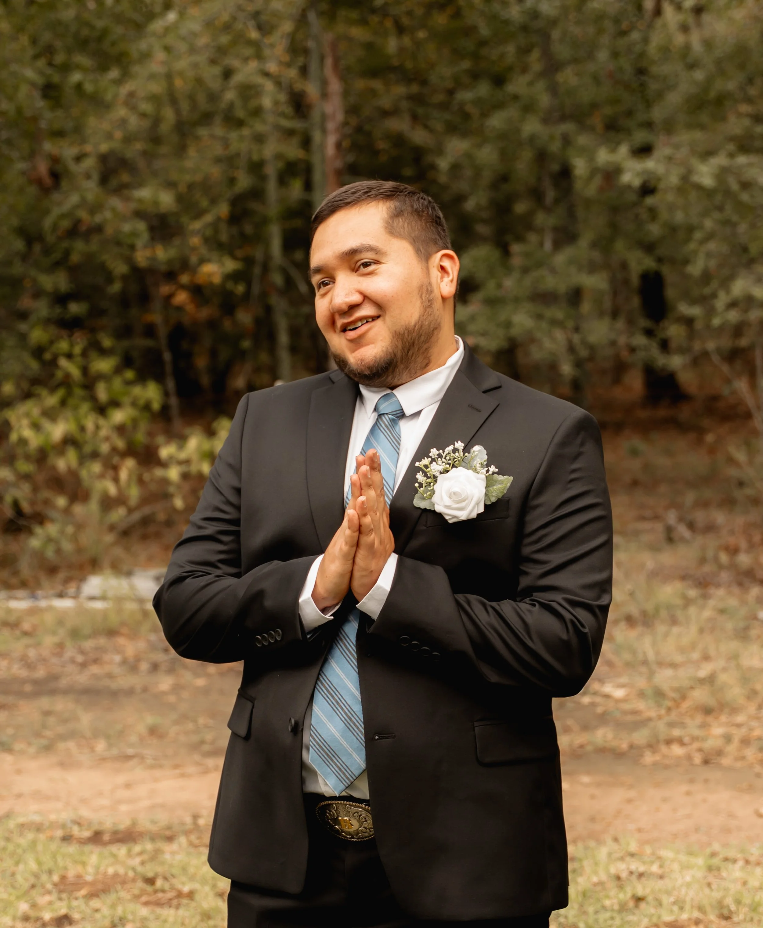 Man in a black suit with a white shirt, striped tie, and boutonniere standing outdoors with hands in prayer position, smiling, with trees and foliage in the background.