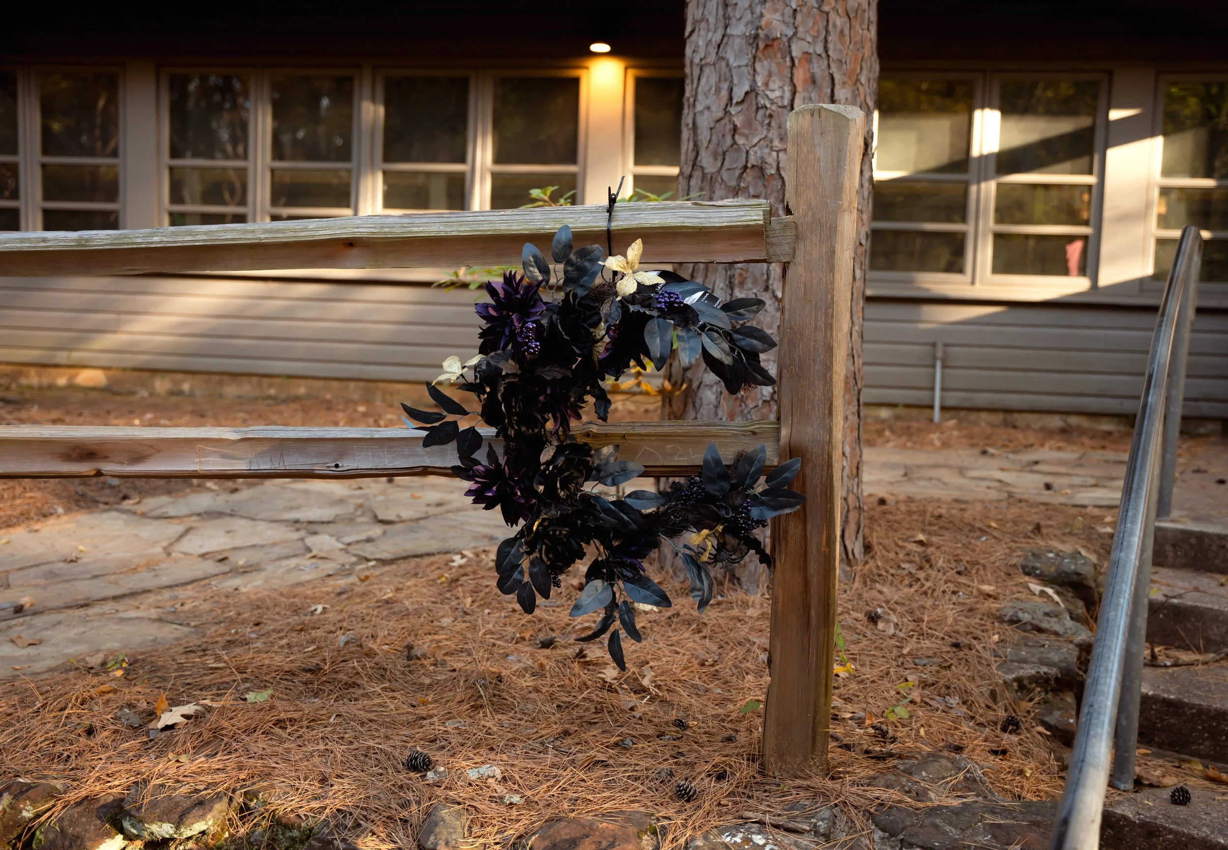 A black wreath hanging on a wooden railing outside near a tree, with a building and windows in the background at sunset.