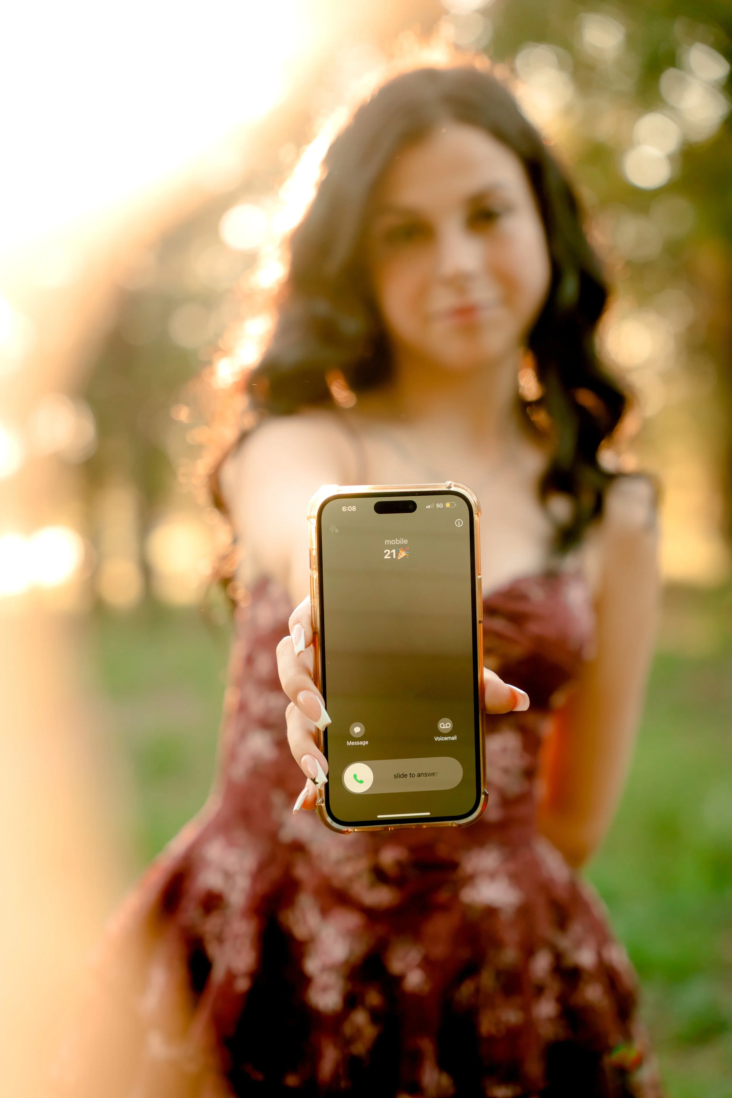 A woman with dark hair in a floral dress holds a smartphone towards the camera, with sunlight in the background.
