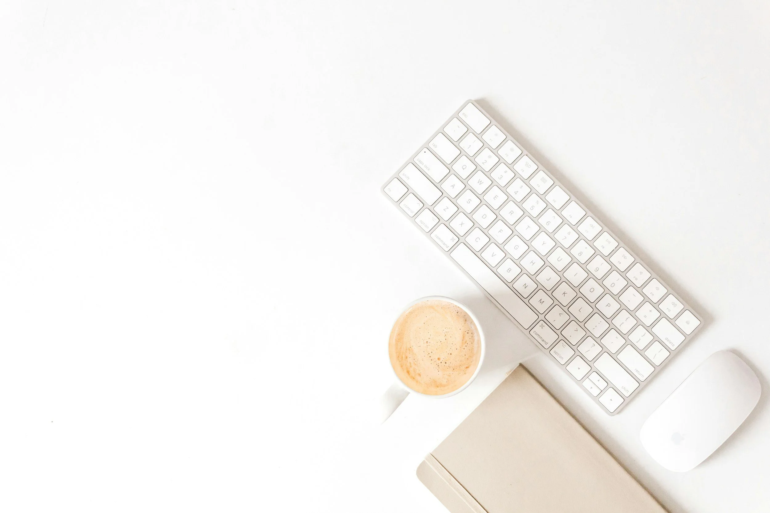 White keyboard and mouse on a desk with a cup of coffee and a closed beige notebook.