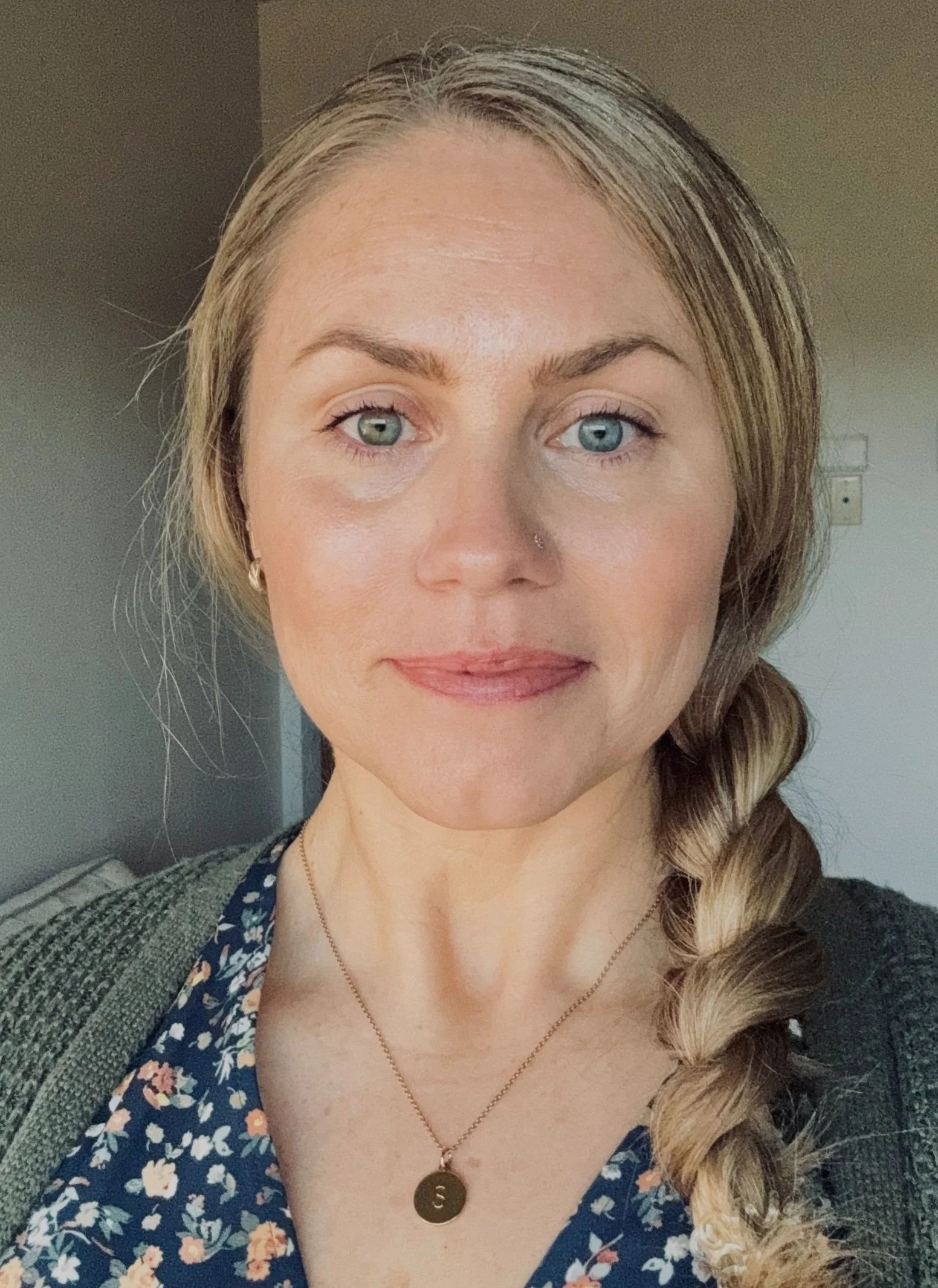 A woman with blonde hair in a braid, blue eyes, wearing a floral blouse, a gold necklace with a pendant, and a green cardigan, standing indoors with a neutral background.