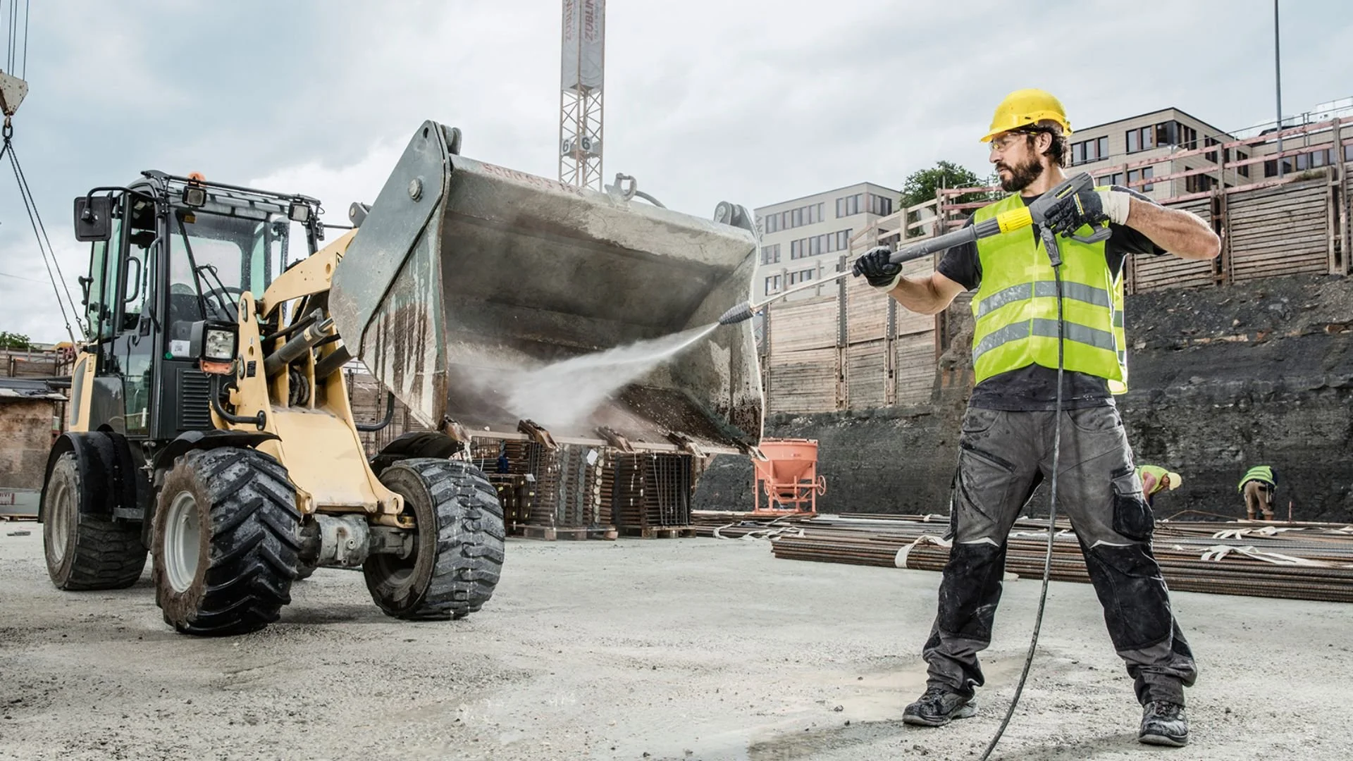 Construction worker using pressure washer on construction site with excavator and workers in background.