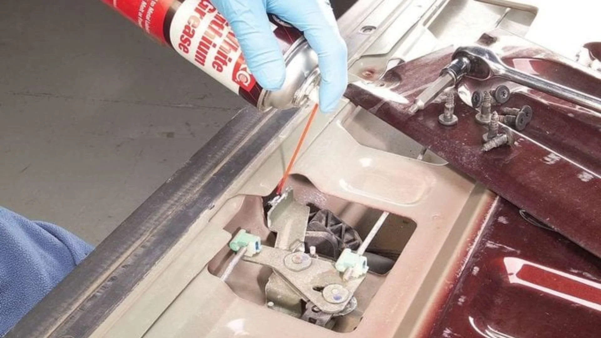 A person wearing blue gloves is using a spray can to apply lubricant or adhesive to a metal part inside a machine or vehicle for maintenance or repair.