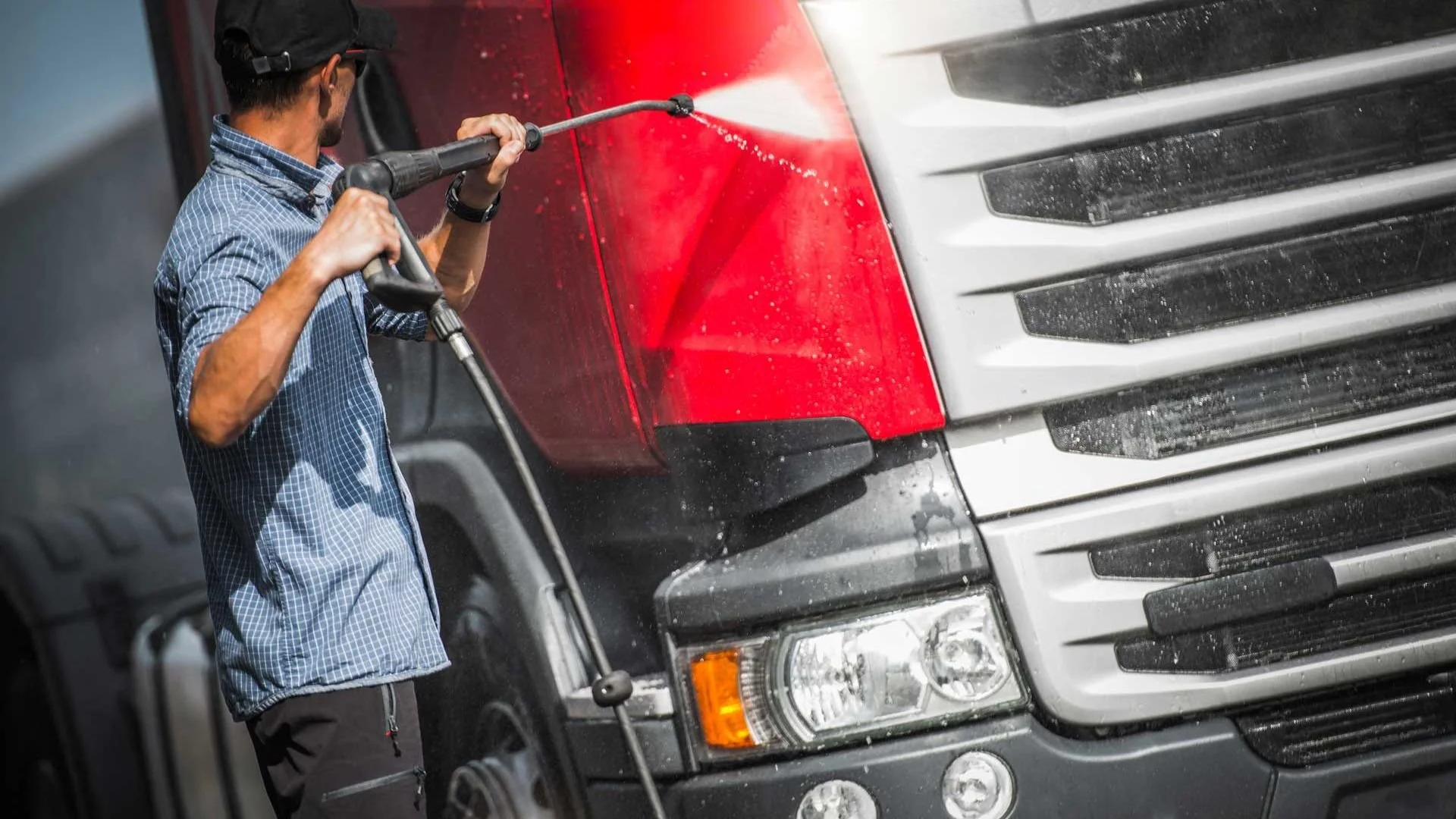 A person wearing a blue checkered shirt and a black cap is pressure washing the front of a large truck with a red and silver exterior.