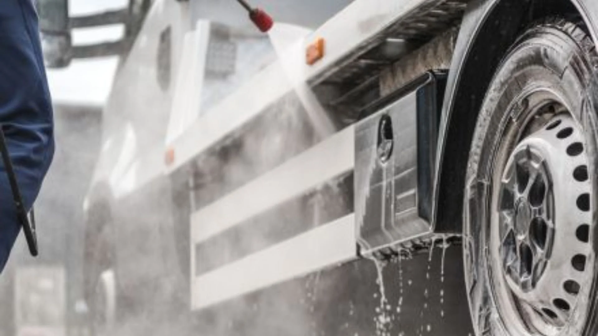 A person is pressure washing the tire and wheel of a vehicle, with water spraying and dripping off the wheel.
