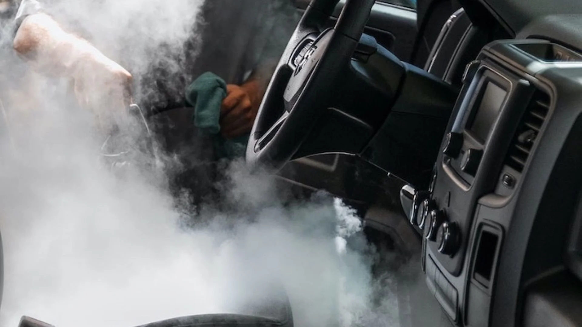 A person is cleaning the dashboard of a vehicle with a green cloth, surrounded by a large amount of smoke or steam.