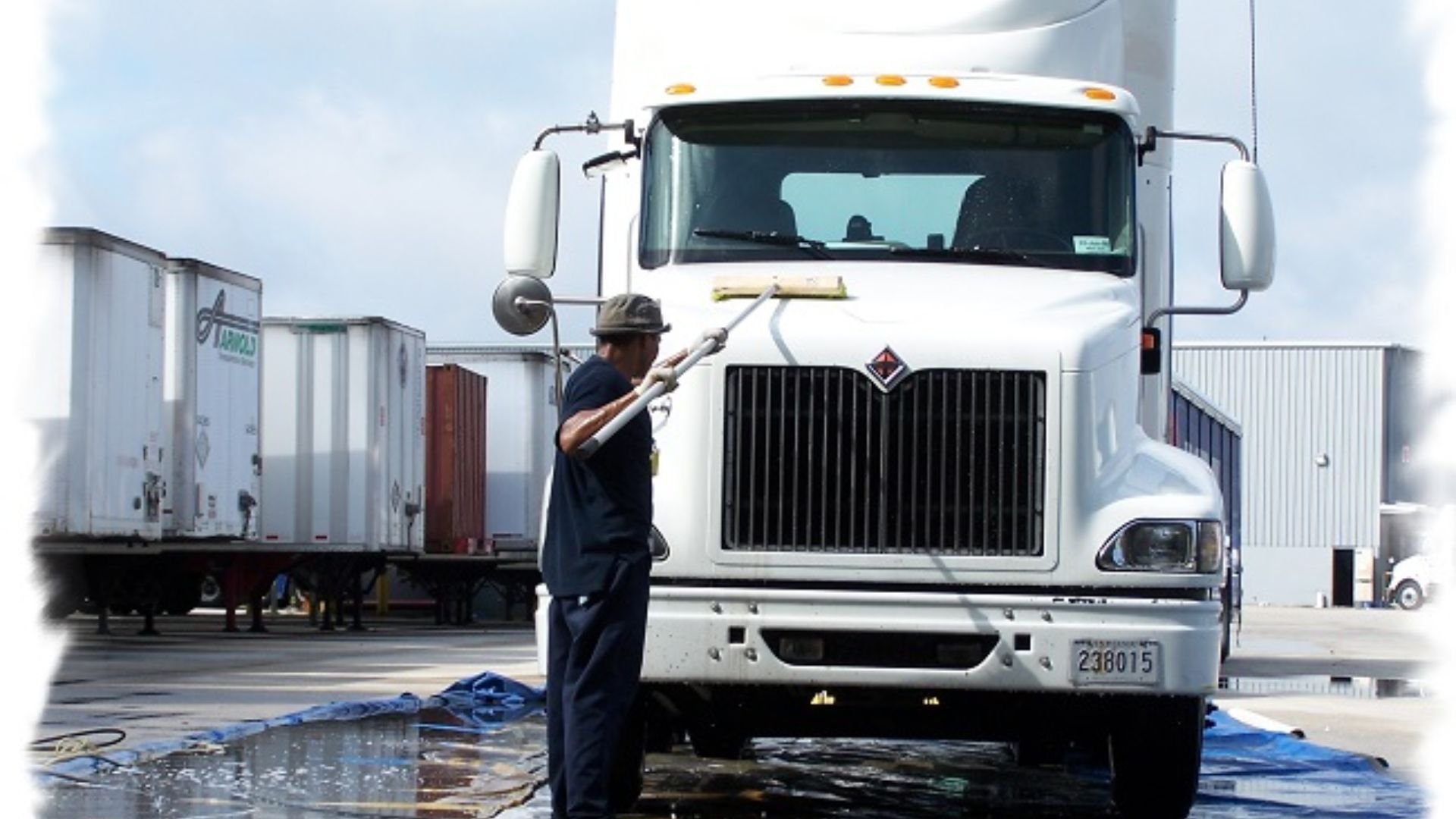 A person cleaning the front of a large white semi-truck with a squeegee, on a truck wash pad with puddles of water and other trucks in the background.