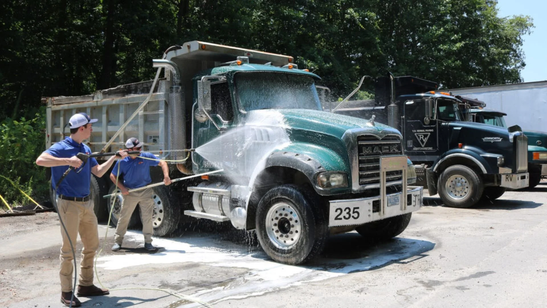 Two men spraying water on a large green dump truck with hoses, while another black truck is parked behind.