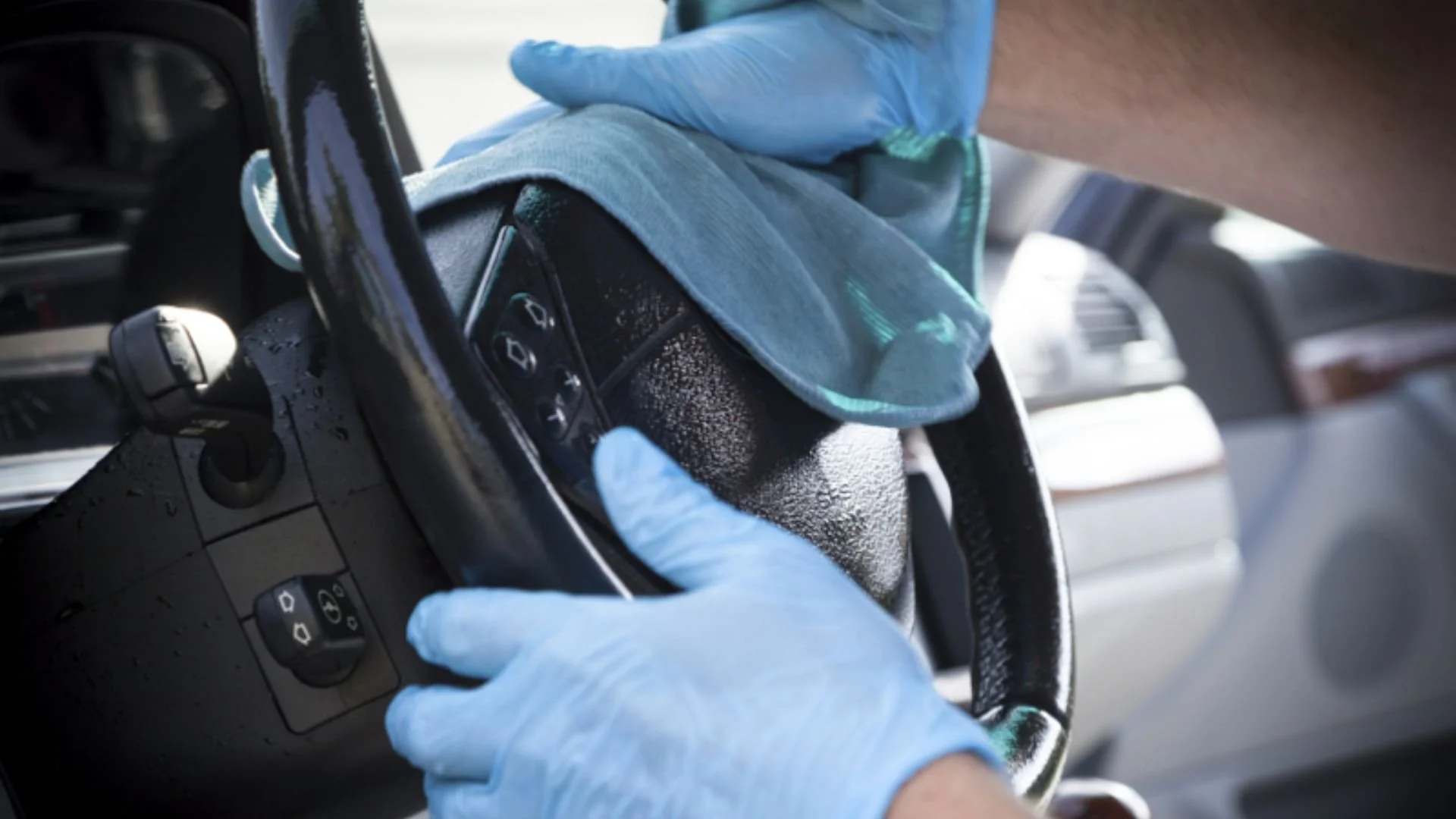 A person wearing blue gloves cleaning the steering wheel of a car with a cloth.