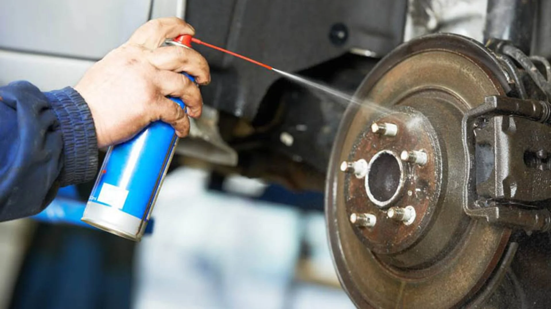 A person sprays brake cleaner onto a rusty car brake rotors in a garage or workshop.