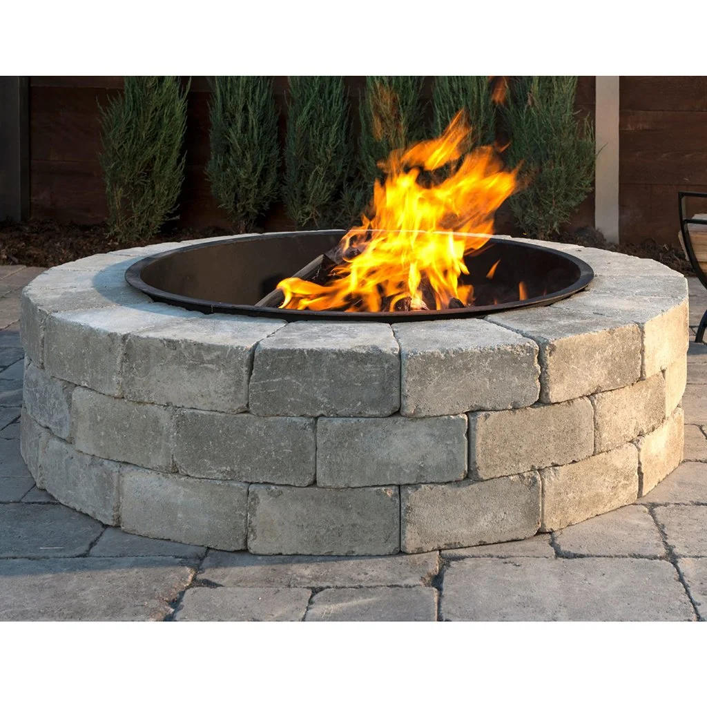 A fire pit built with stacked stone blocks with a metal basin in the center, burning with bright flames, outdoors on a paved patio with green shrubs in the background.