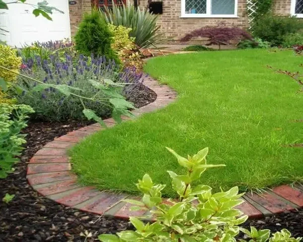 Curved brick pathway in a garden with green grass, flowering plants, and shrubs.